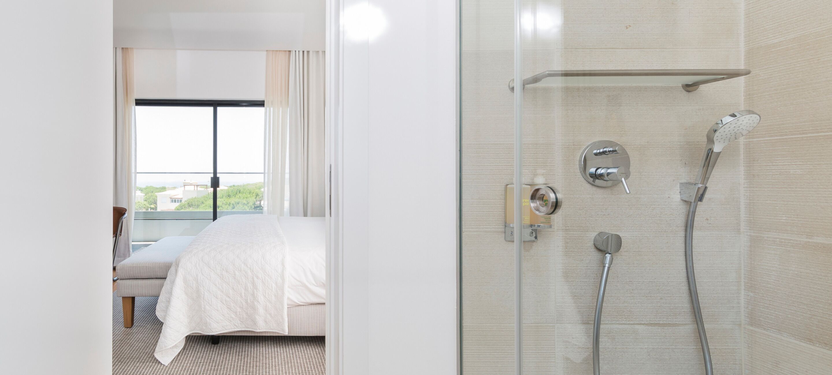 A hotel room with a bed, a shower with a glass door, and a view of greenery outside.