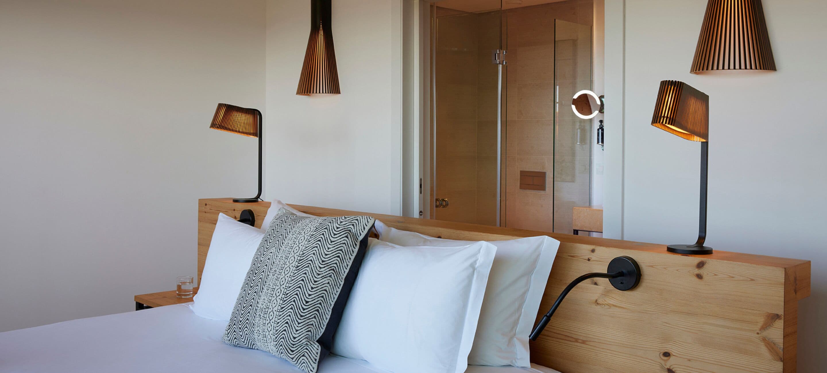 A modern hotel room with a wooden headboard, a bed with white and patterned pillows, and two bedside lamps.