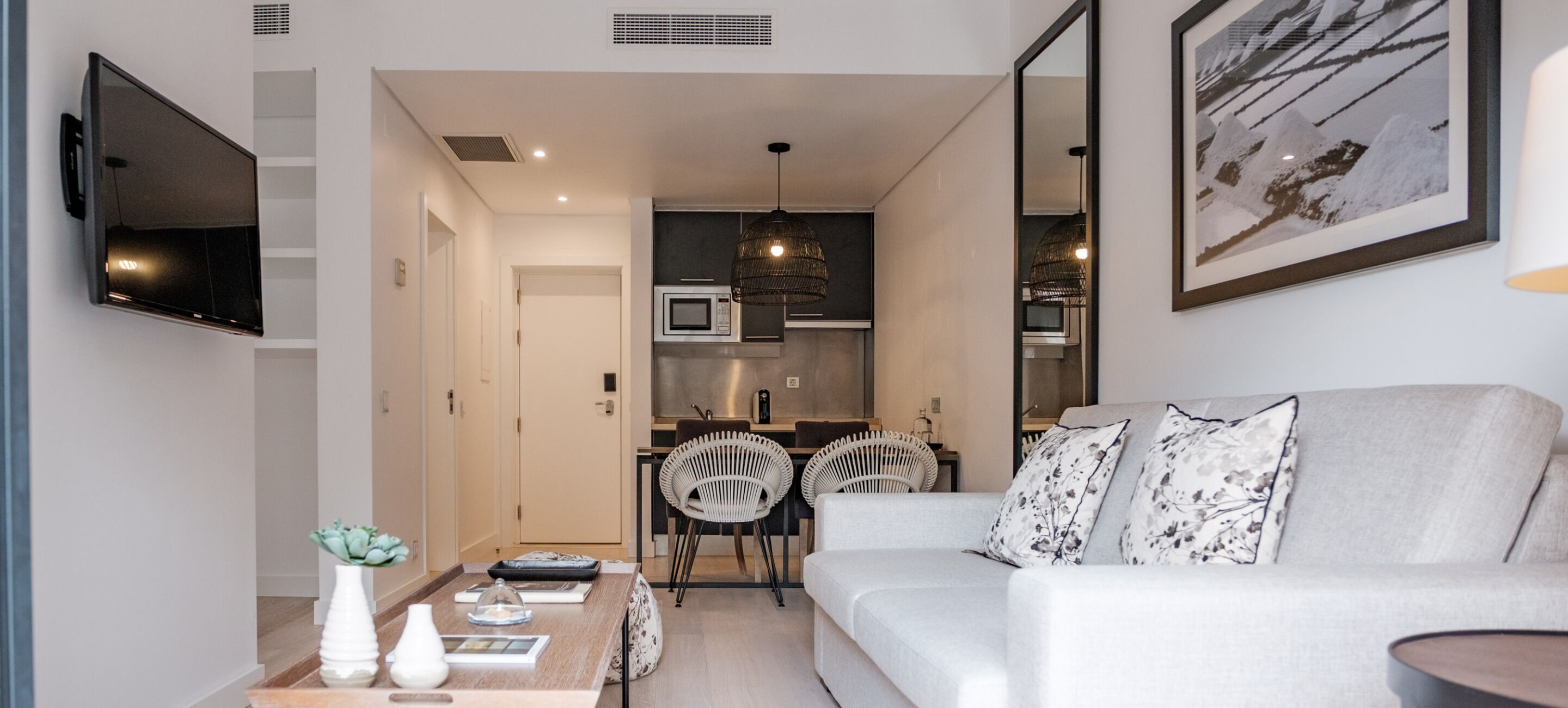 A modern living room with a sofa, a coffee table, and a TV mounted on the wall. There is a kitchen area with a microwave and a dining table with chairs.