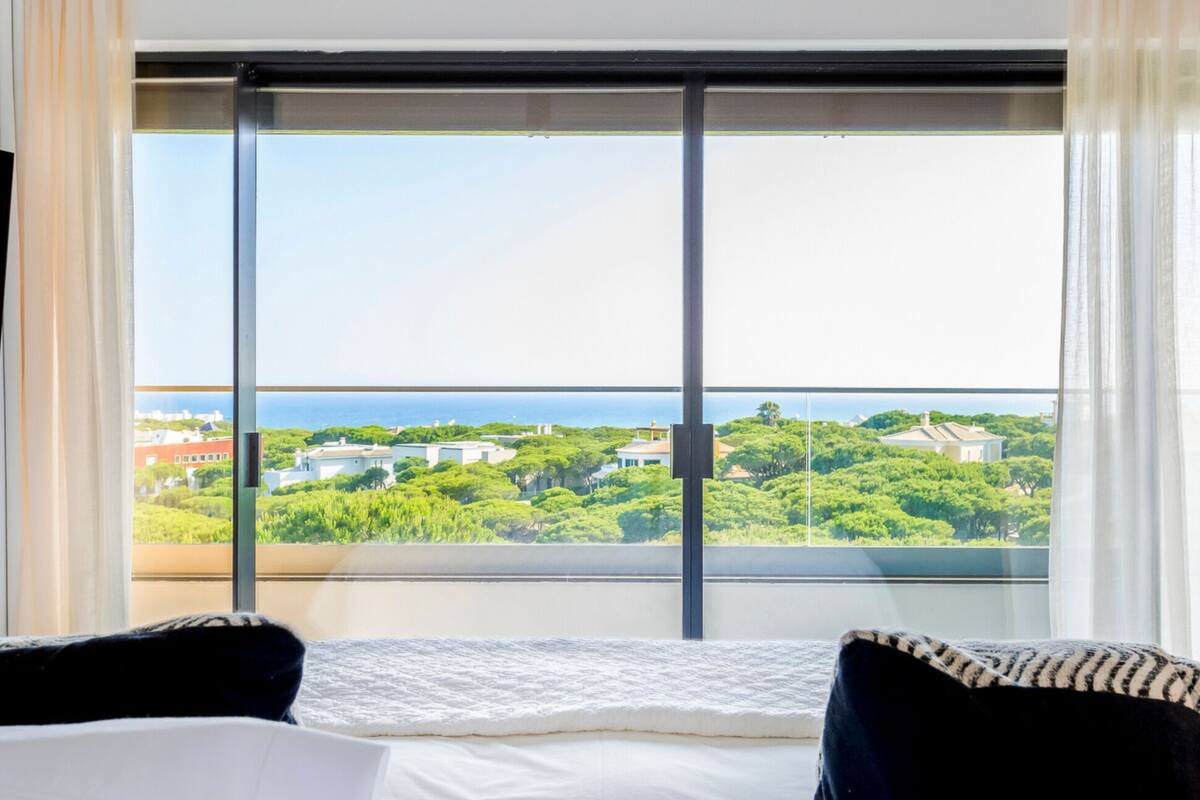 A hotel room with a large window, a bed with white bedding, and a view of a green landscape and buildings.