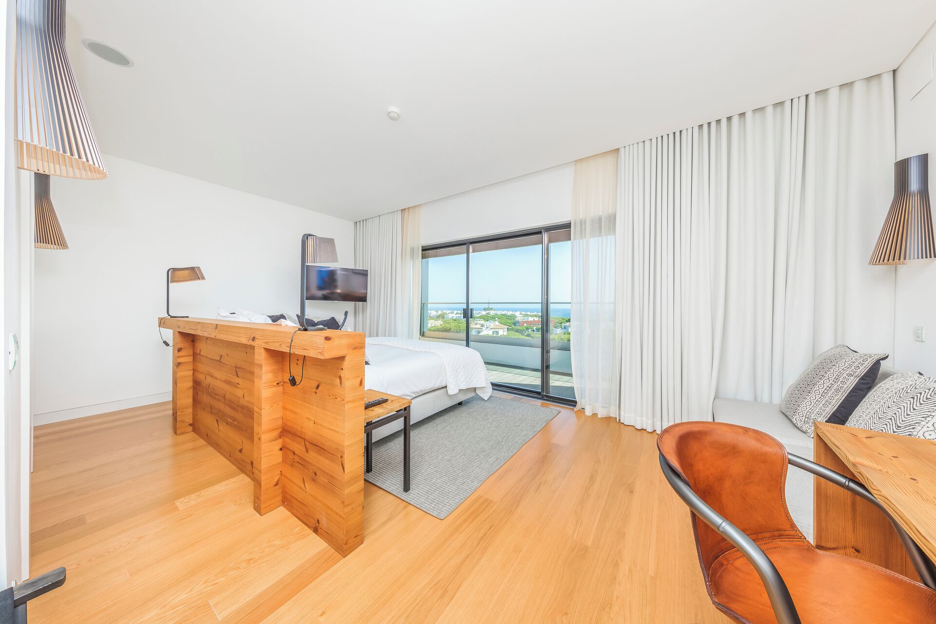 A modern hotel room with a wooden desk, a bed, a TV, and a view of the city.