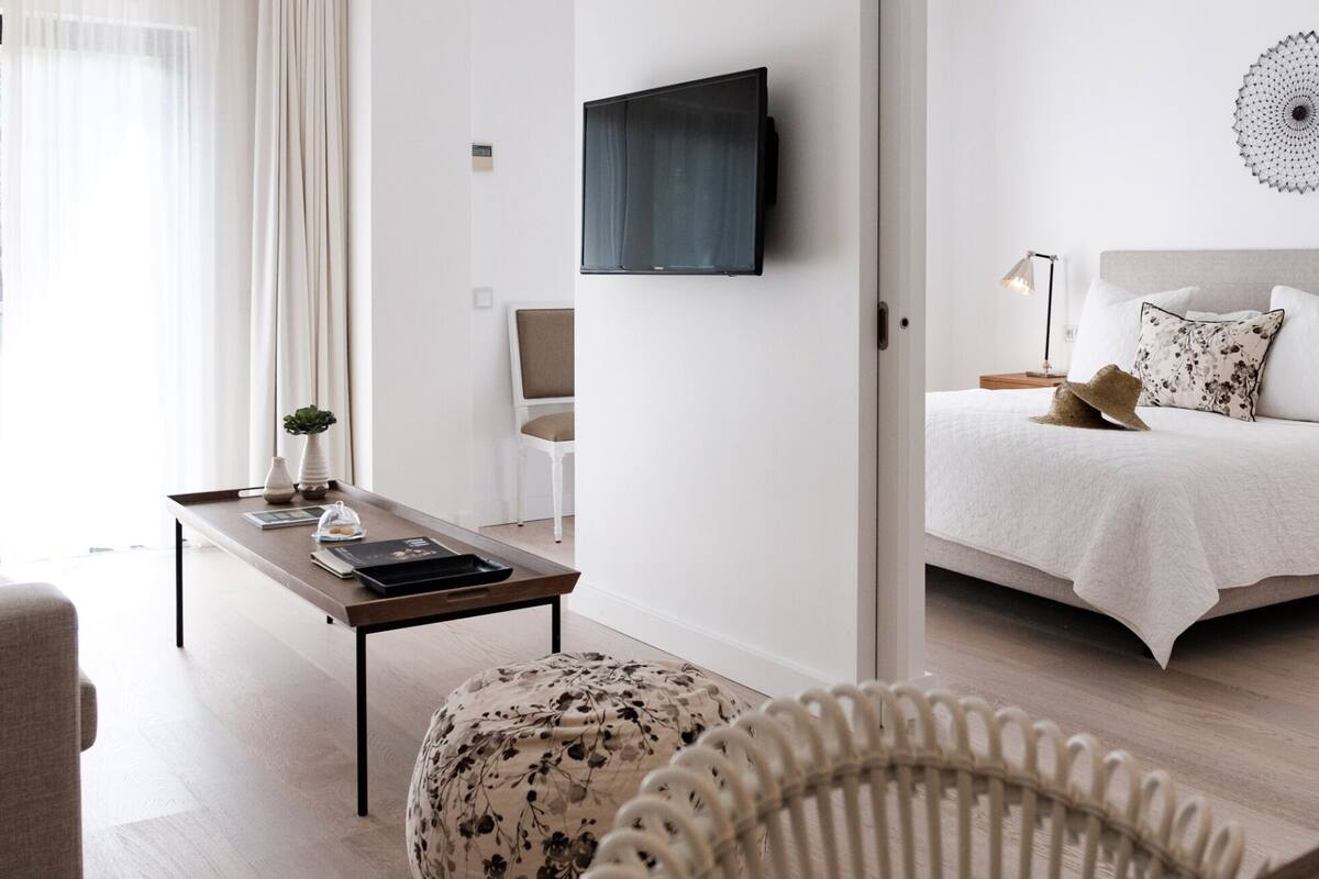 A modern hotel room with a bed, a TV mounted on the wall, a small table with a lamp, and a window with sheer curtains.