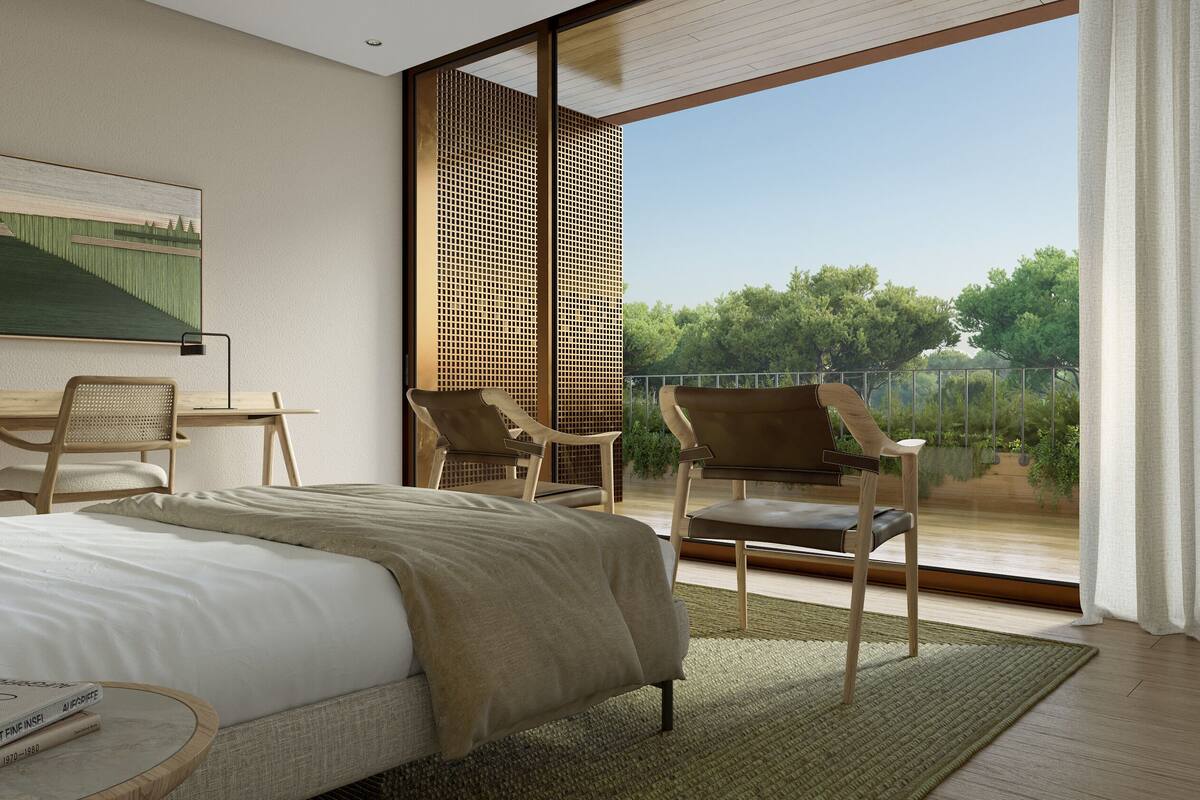 A modern bedroom with a large bed, a desk, two chairs, and a balcony with a view of trees.