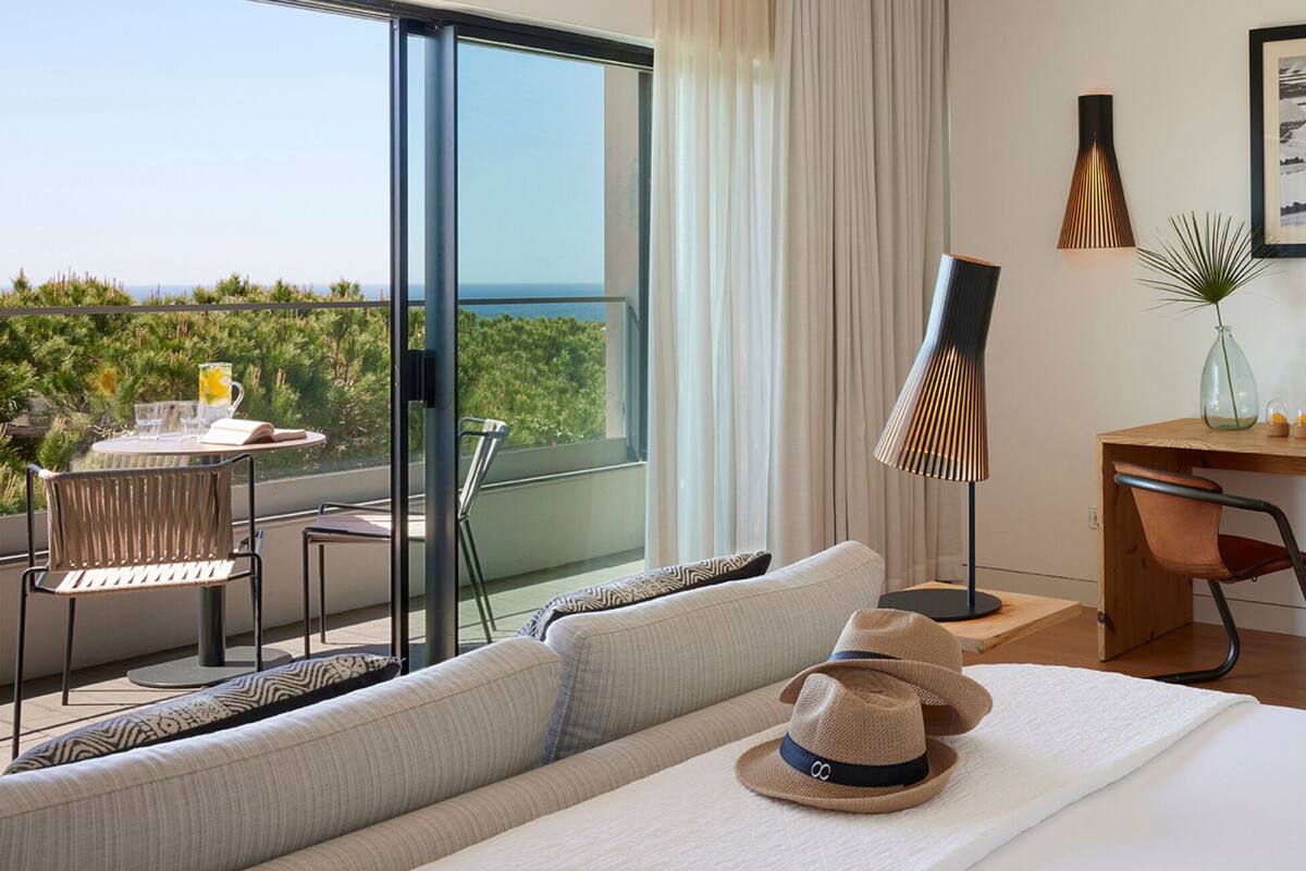 A modern hotel room with a balcony, a dining area, a desk, and a large window offering a scenic view.