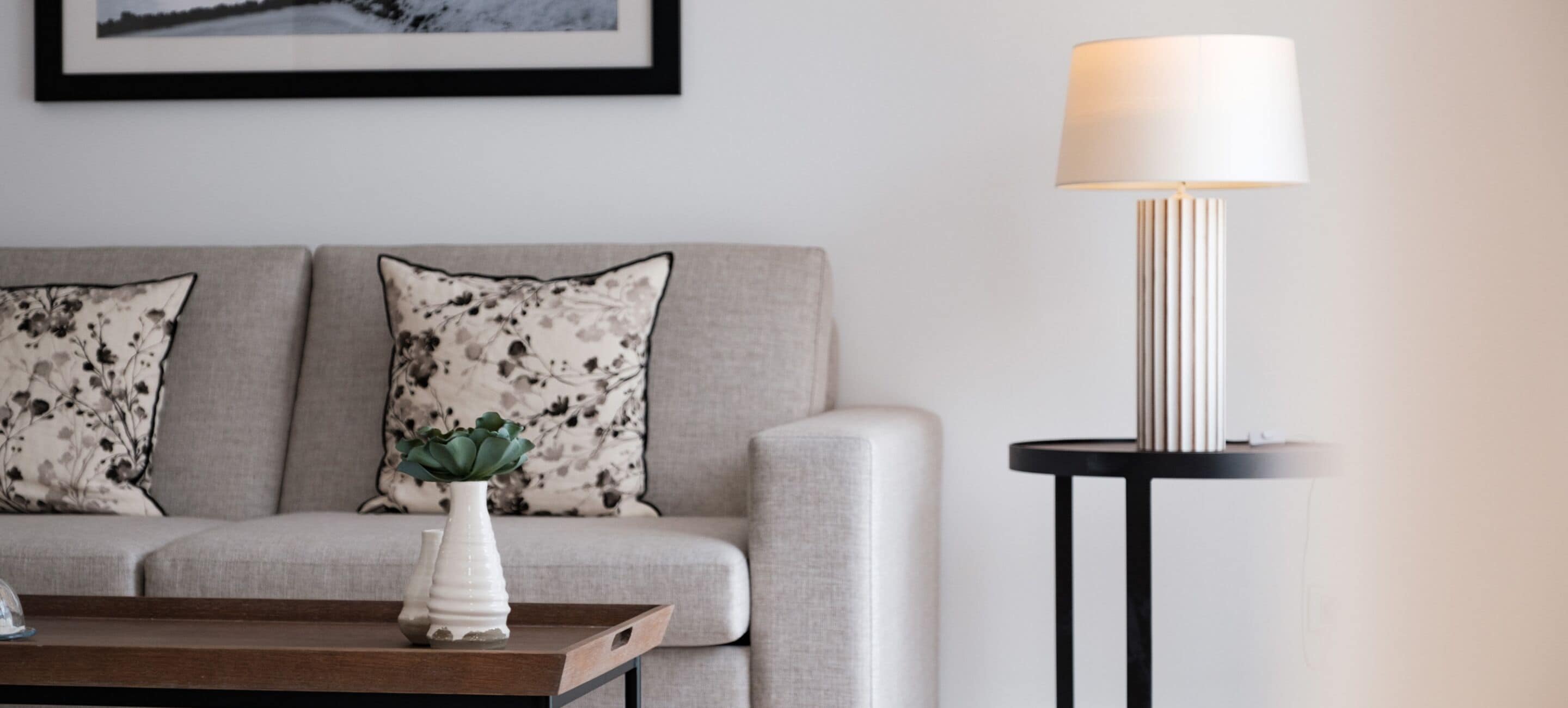 Una sala de estar moderna con un sofá gris, cojines estampados, un jarrón blanco con una planta y una lámpara de pie alta.