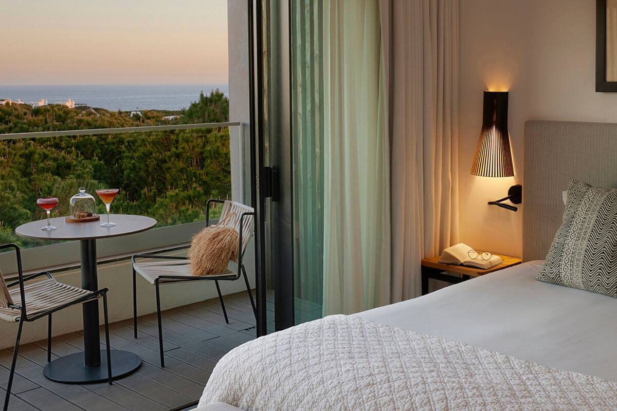 A hotel room with a balcony, a bed with patterned pillows, a table with two drinks, and a view of the sea.