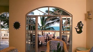 Eine sonnendurchflutete Terrasse mit Korbmöbeln, eine Glastür zu einem Essbereich mit Tischen und ein dekoratives Meeresjungfern-Wandgemälde.
