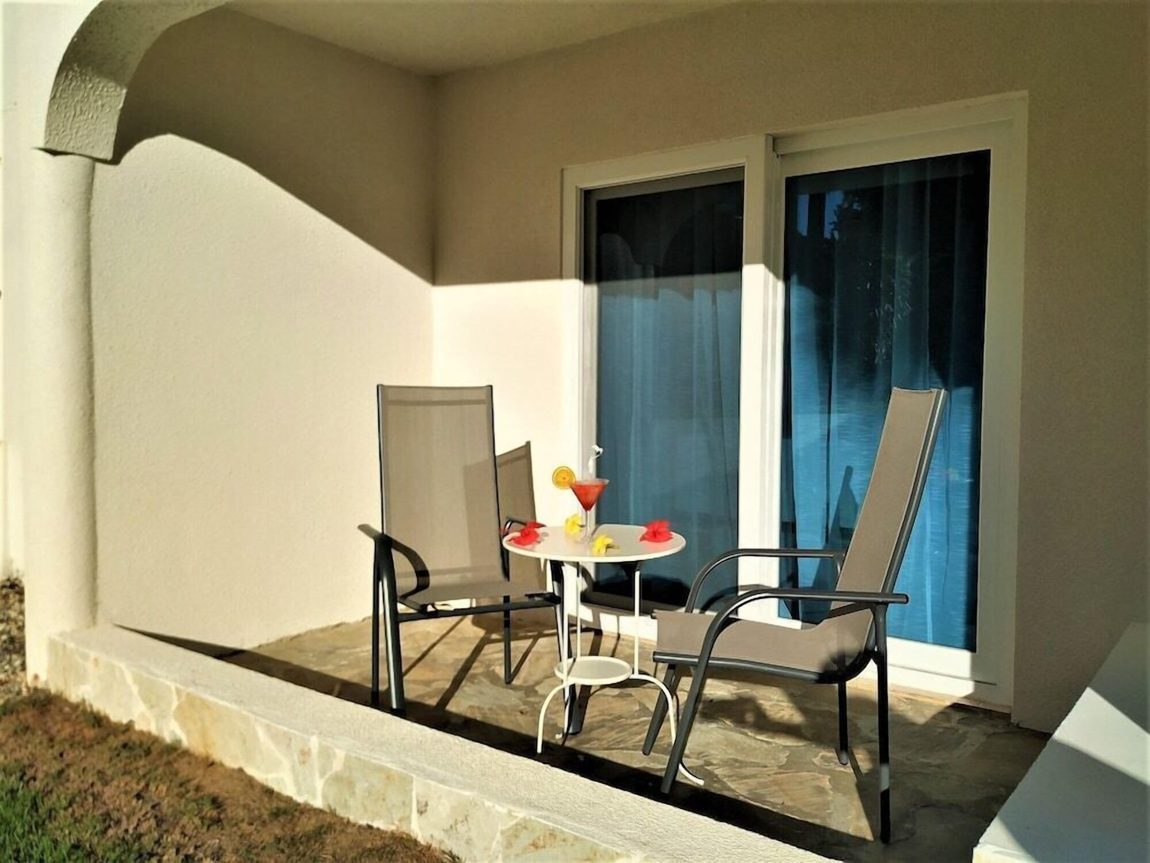 Uma varanda com duas cadeiras e uma pequena mesa, uma janela com cortinas azuis e um parapeito de pedra.