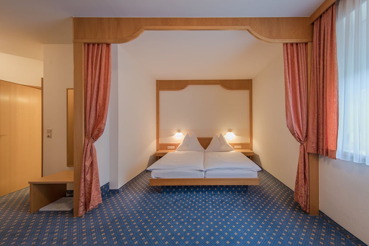 A hotel room with a bed, two bedside tables, a nightstand, a wooden headboard, and a window with curtains.