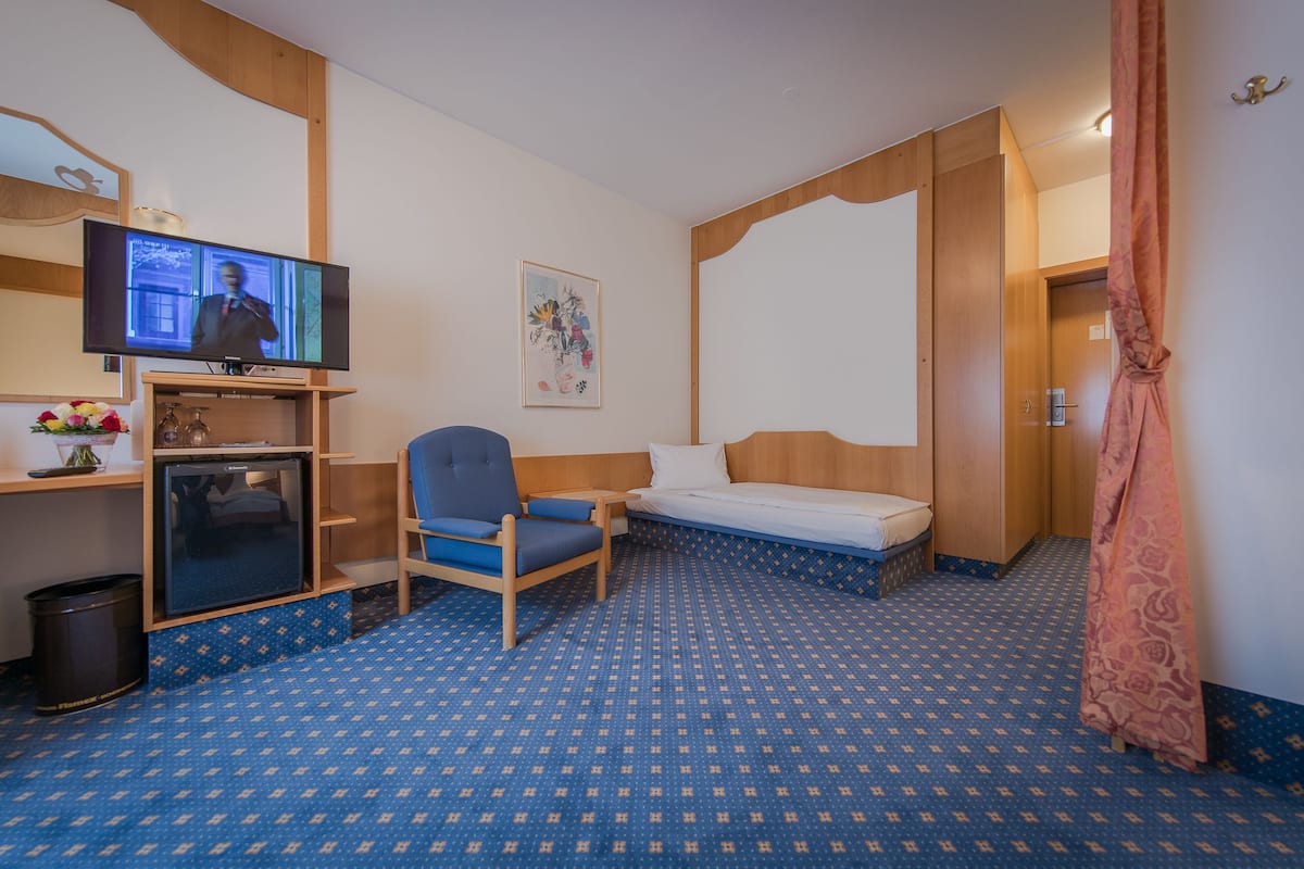 A hotel room with a bed, a desk with a television, a chair, a small refrigerator, and a trash bin.