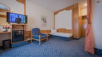 A hotel room with a bed, a desk with a television, a chair, a small refrigerator, and a trash bin.