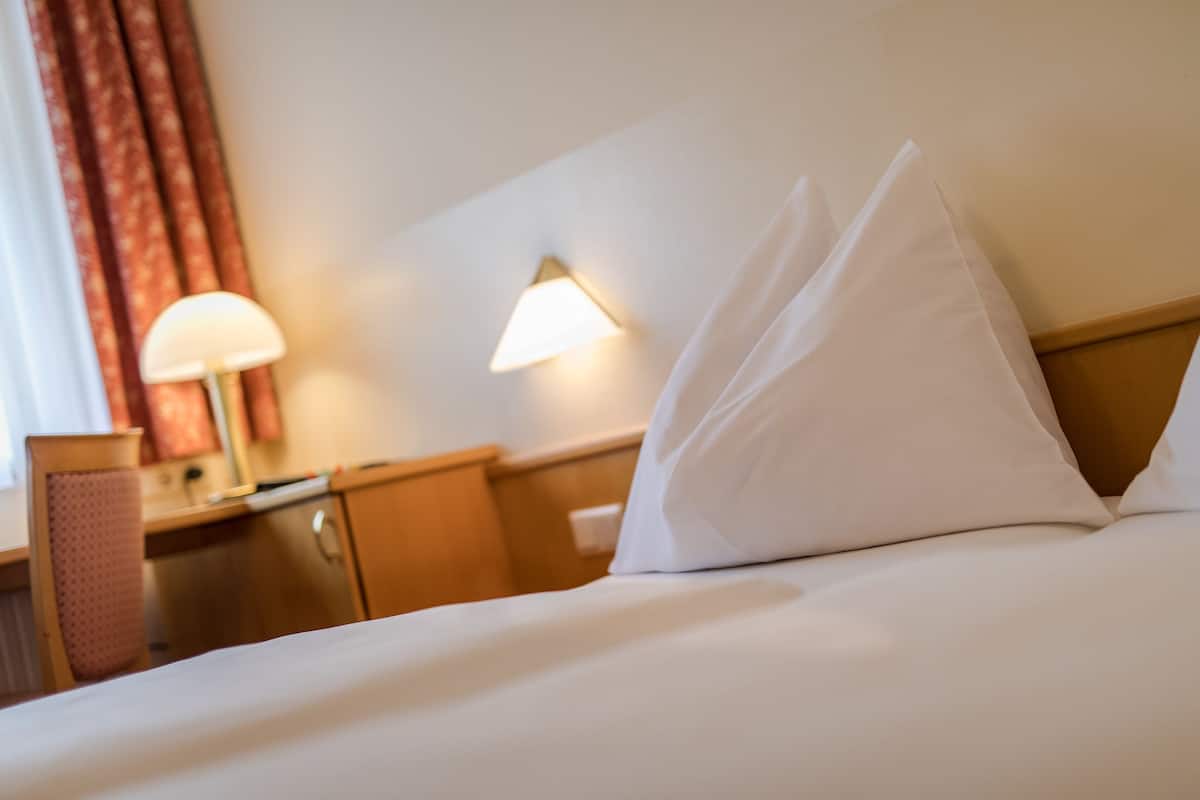 A hotel room with a neatly made bed, a lamp, a wall-mounted light, and a window with curtains.