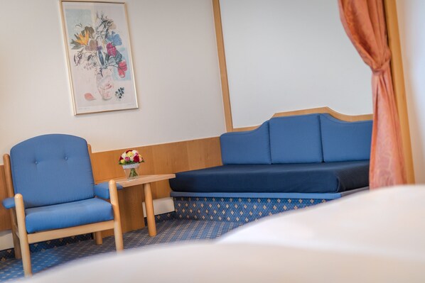 A hotel room with a blue sofa, a blue armchair, a small table with a vase of flowers, and a framed artwork on the wall.