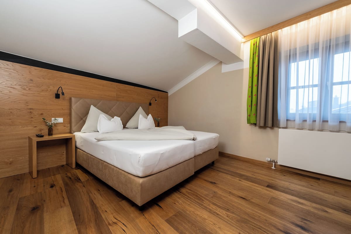 Een moderne slaapkamer met een houten vloer, een bed met witte lakens, een nachtkastje, een wandlamp en een raam met dunne gordijnen.