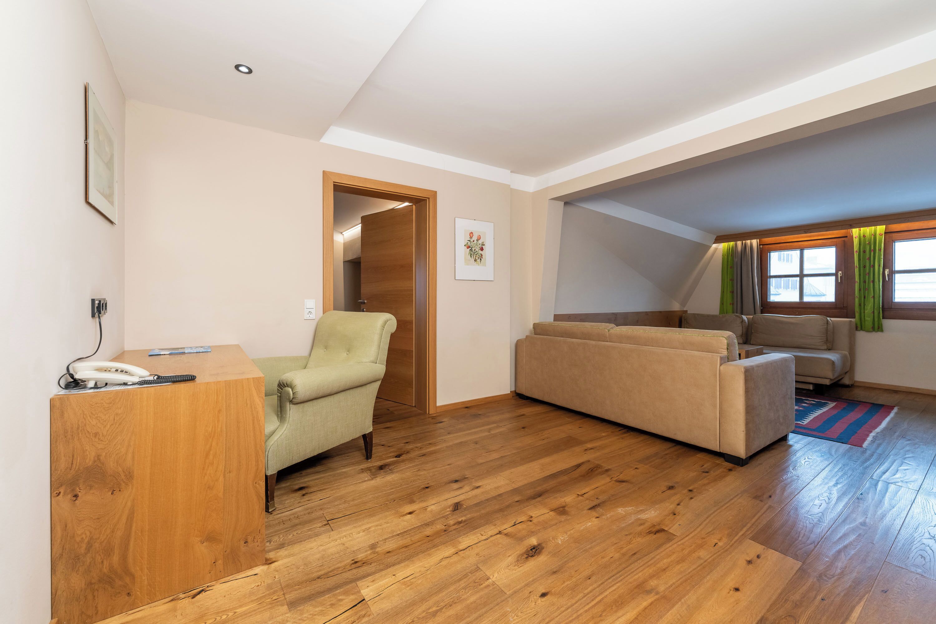 A spacious living area with a wooden floor, a green armchair, a beige sofa, and a wooden desk with a telephone.