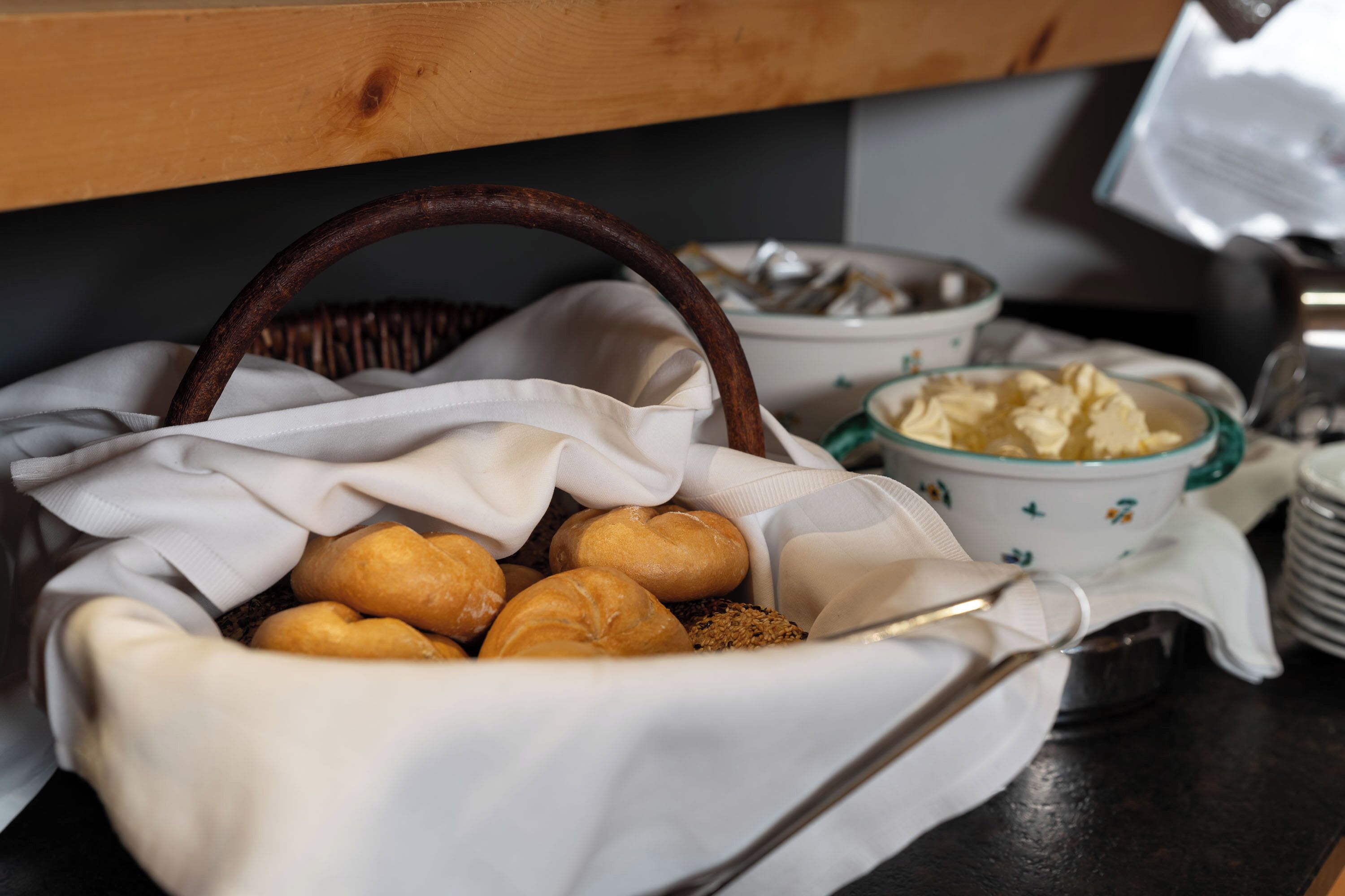 Un panier de viennoiseries est posé sur une table, à côté d’une serviette en tissu, d’un bol de beurre et d’une pile d’assiettes.