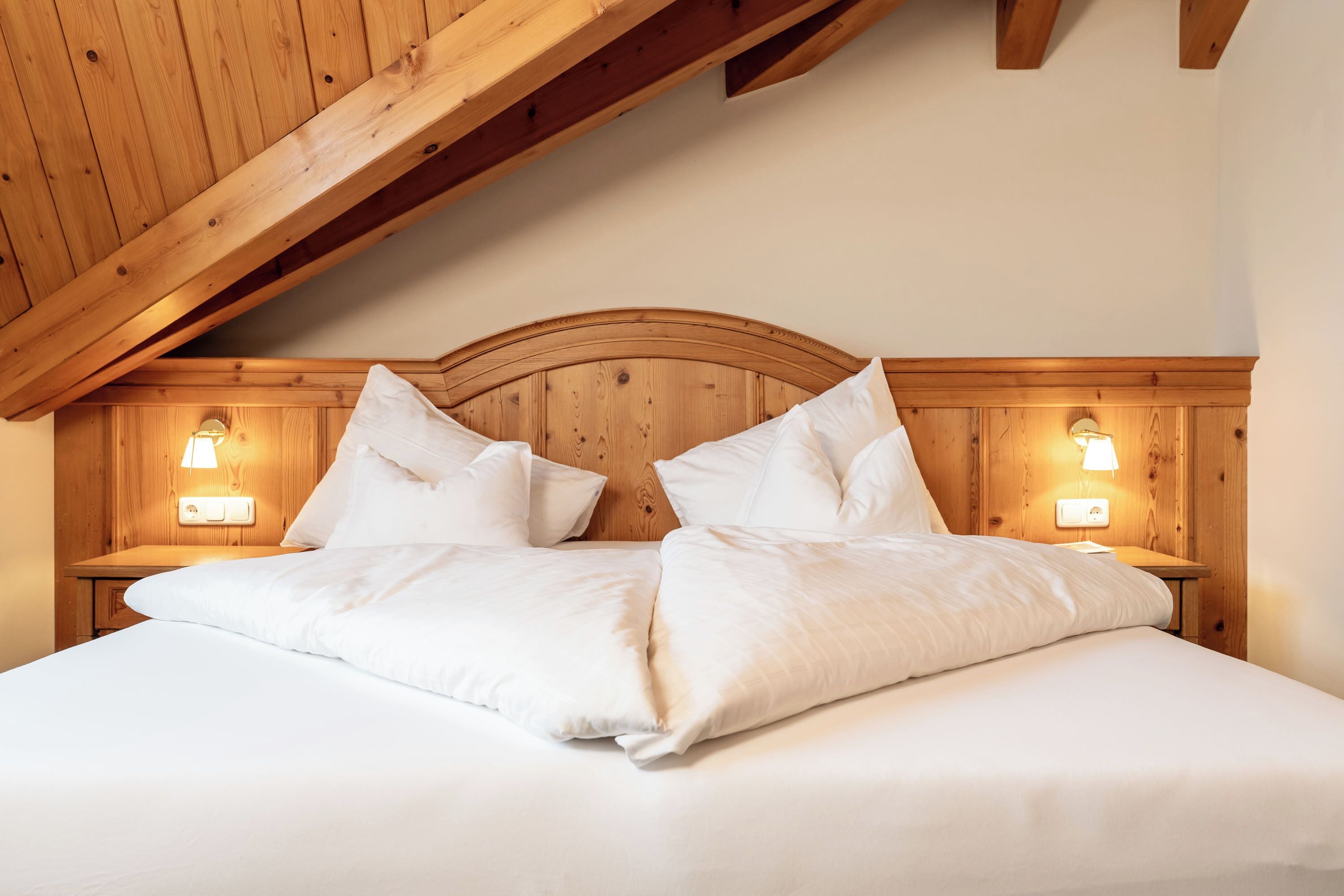 Una cama bien hecha con sábanas blancas y almohadas, cabecera de madera y dos lámparas de noche.