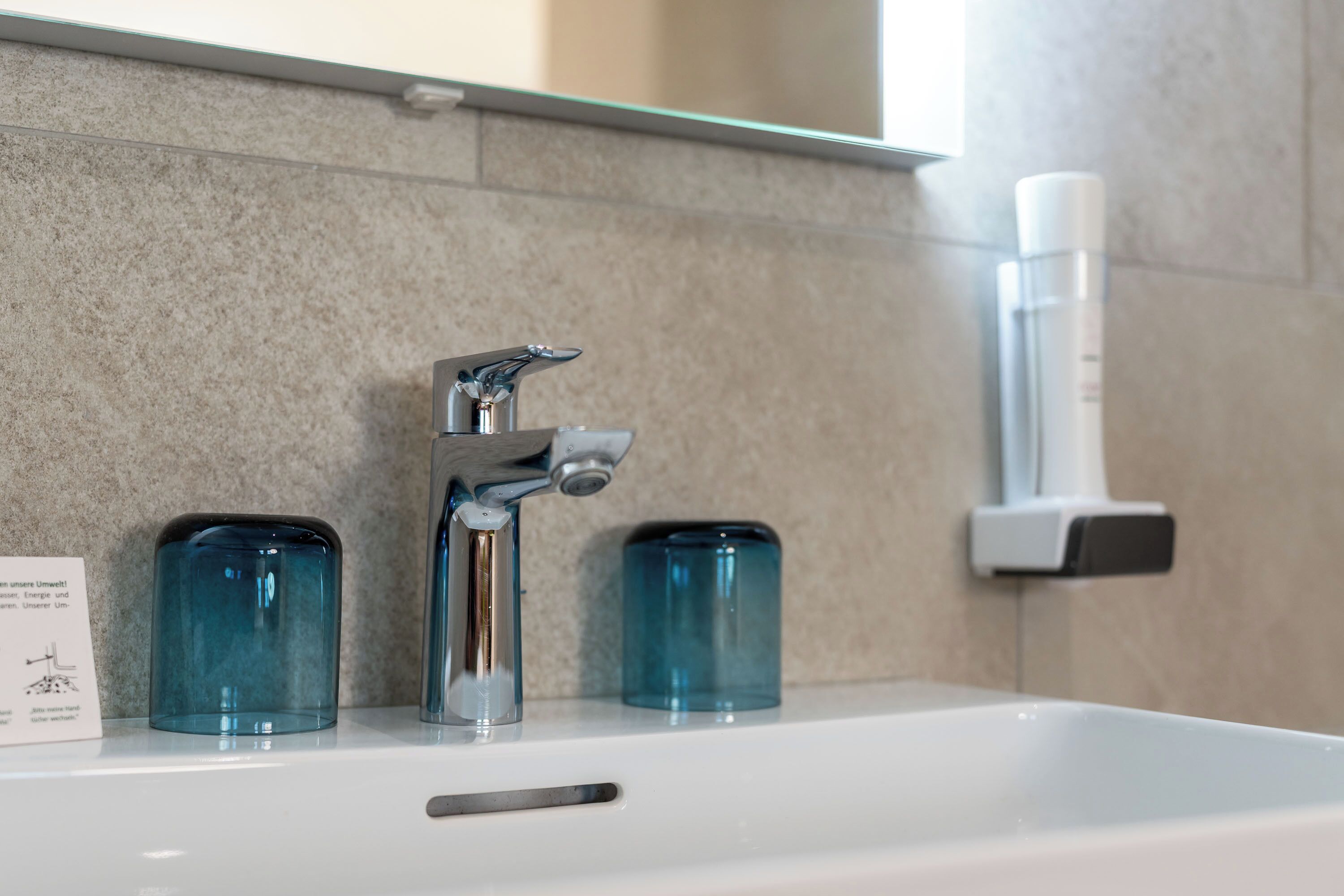 A bathroom sink with a modern faucet, two blue glass containers, and a hand sanitizer dispenser on the wall.