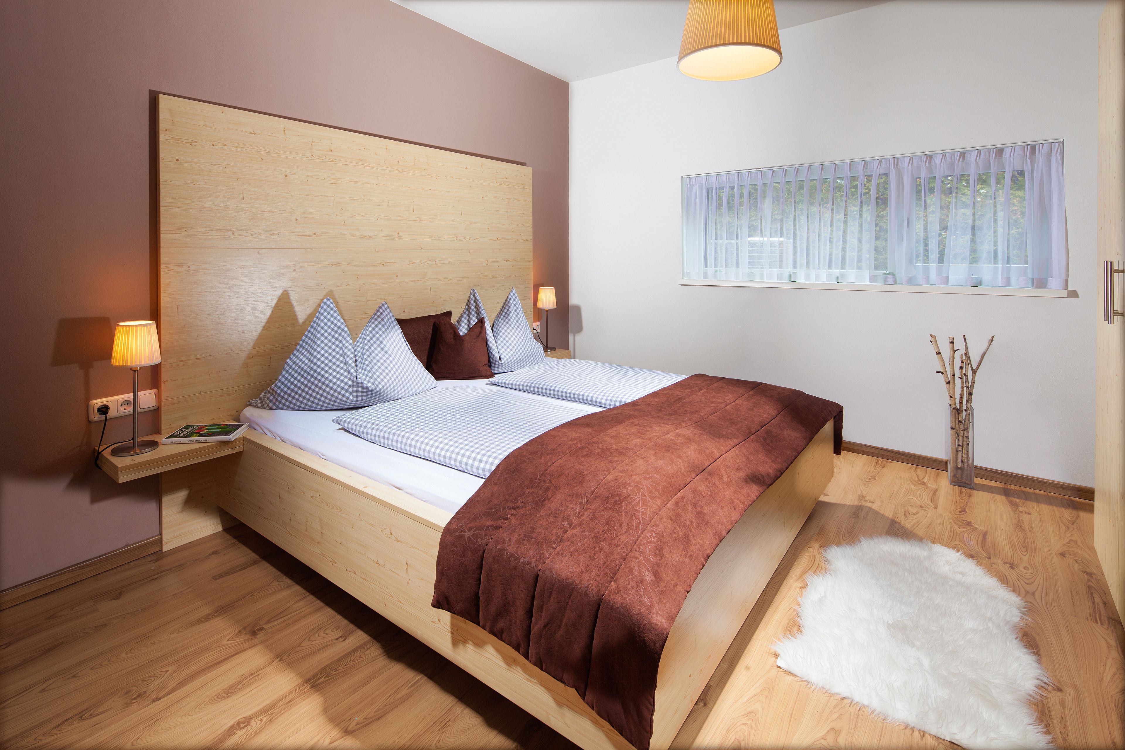 Une chambre moderne avec une tête de lit en bois, un grand lit recouvert d’une couverture marron, deux tables de chevet avec des lampes et une fenêtre ornée de rideaux légers.