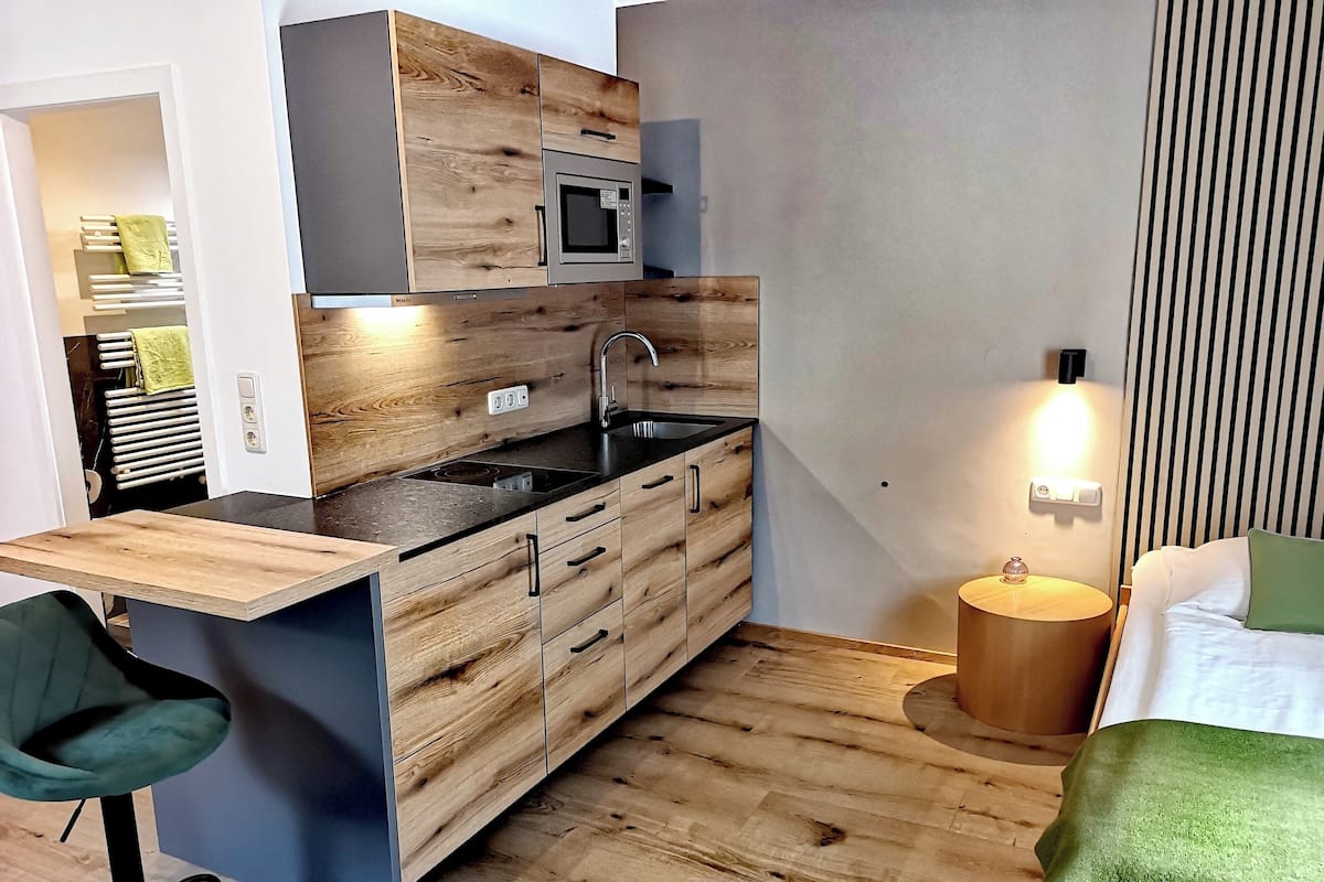 Una cocina compacta con microondas integrado, lavamanos y armarios de madera. Hay una pequeña zona de comedor con una silla y una mesa redonda. La habitación tiene una cama con un cojín verde y una lámpara de noche.
