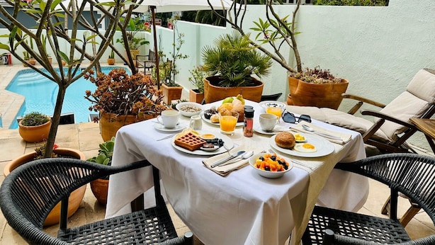 An outdoor dining area with a table set for breakfast, surrounded by potted plants and a pool in the background. - Loloho Lodge (Cape Town)