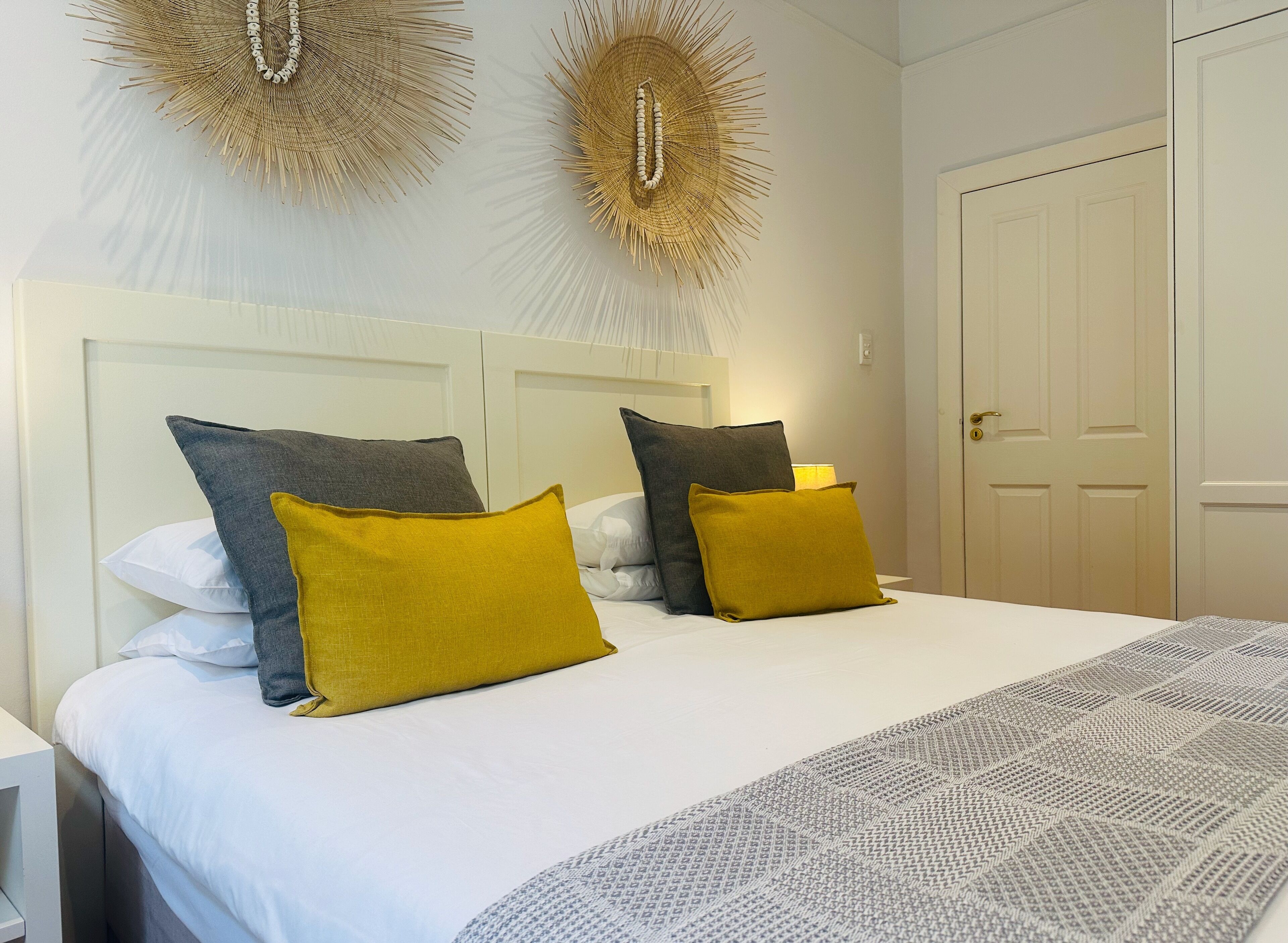 A neatly made bed with white and mustard pillows, a patterned throw, and two wall-mounted decorative items.