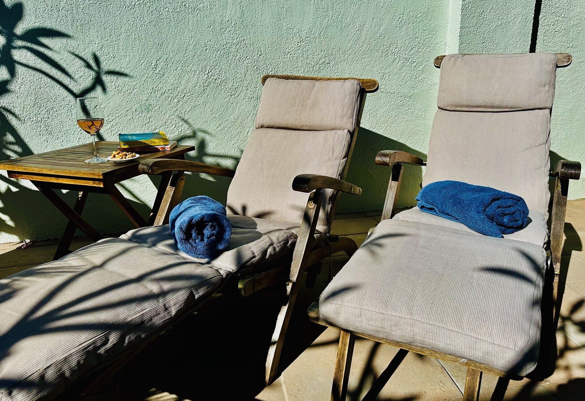 Two wooden lounge chairs with cushions, a small table with a glass and a book, and a blue towel on one of the chairs.