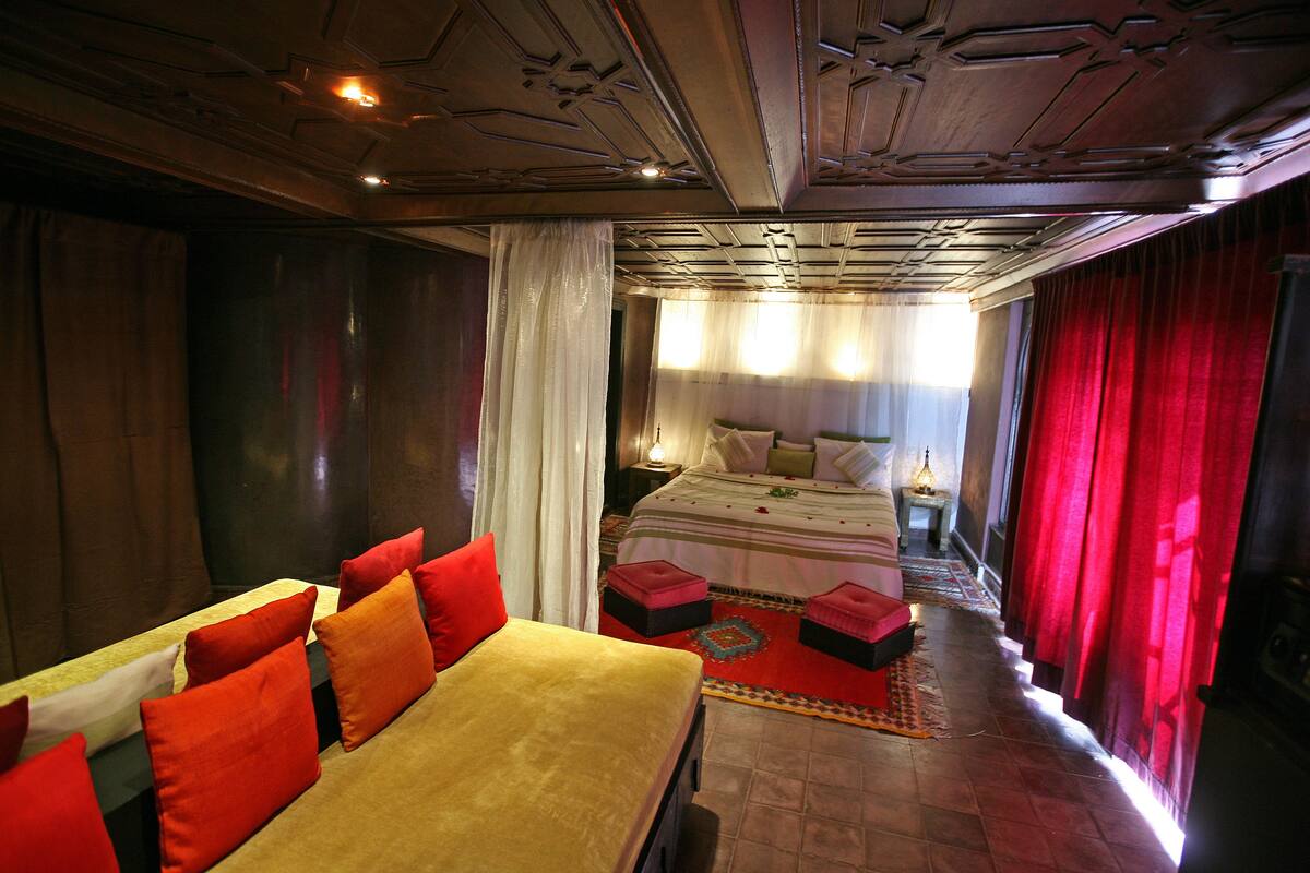 Habitación de hotel con una cama grande, un sofá con cojines rojos, un tapete estampado y techo de madera.