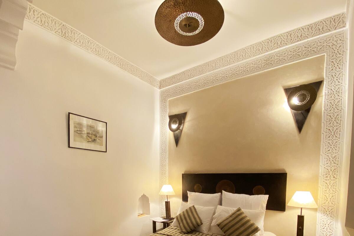 Un dormitorio ordenado con una cama grande, dos mesitas de noche con lámparas, una lámpara decorativa en el techo y un cuadro enmarcado en la pared.