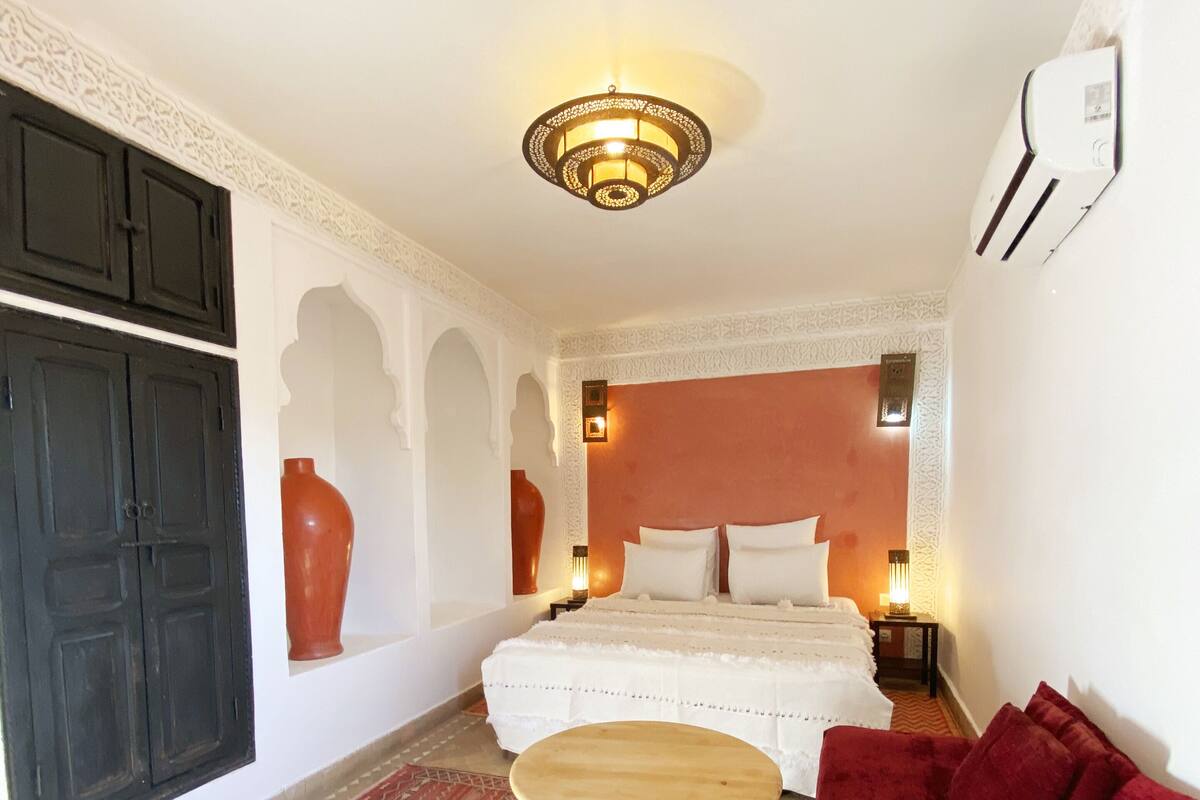 Un dormitorio con cama, sofá rojo, mesa de madera, alfombras estampadas y una lámpara decorativa en el techo.