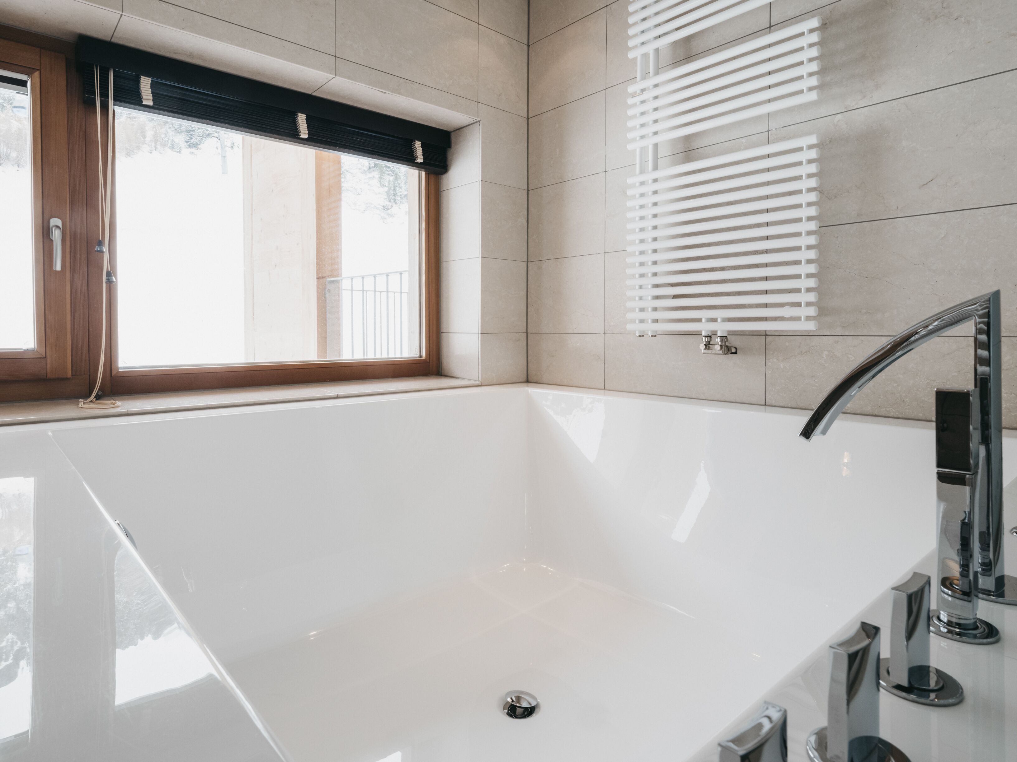 A modern bathroom with a large white bathtub, a window with blinds, and a wall-mounted towel rack.