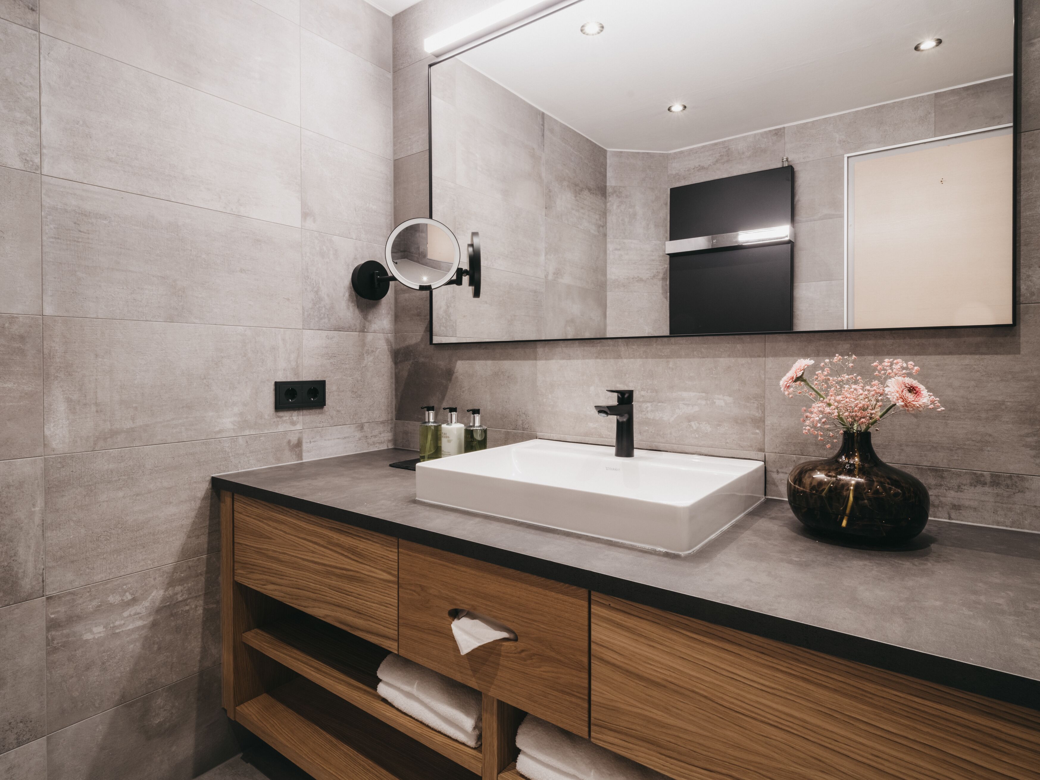 Ein modernes Badezimmer mit einem großen Spiegel, einem weißen Waschbecken, Holzschränken und einer Vase mit Blumen.