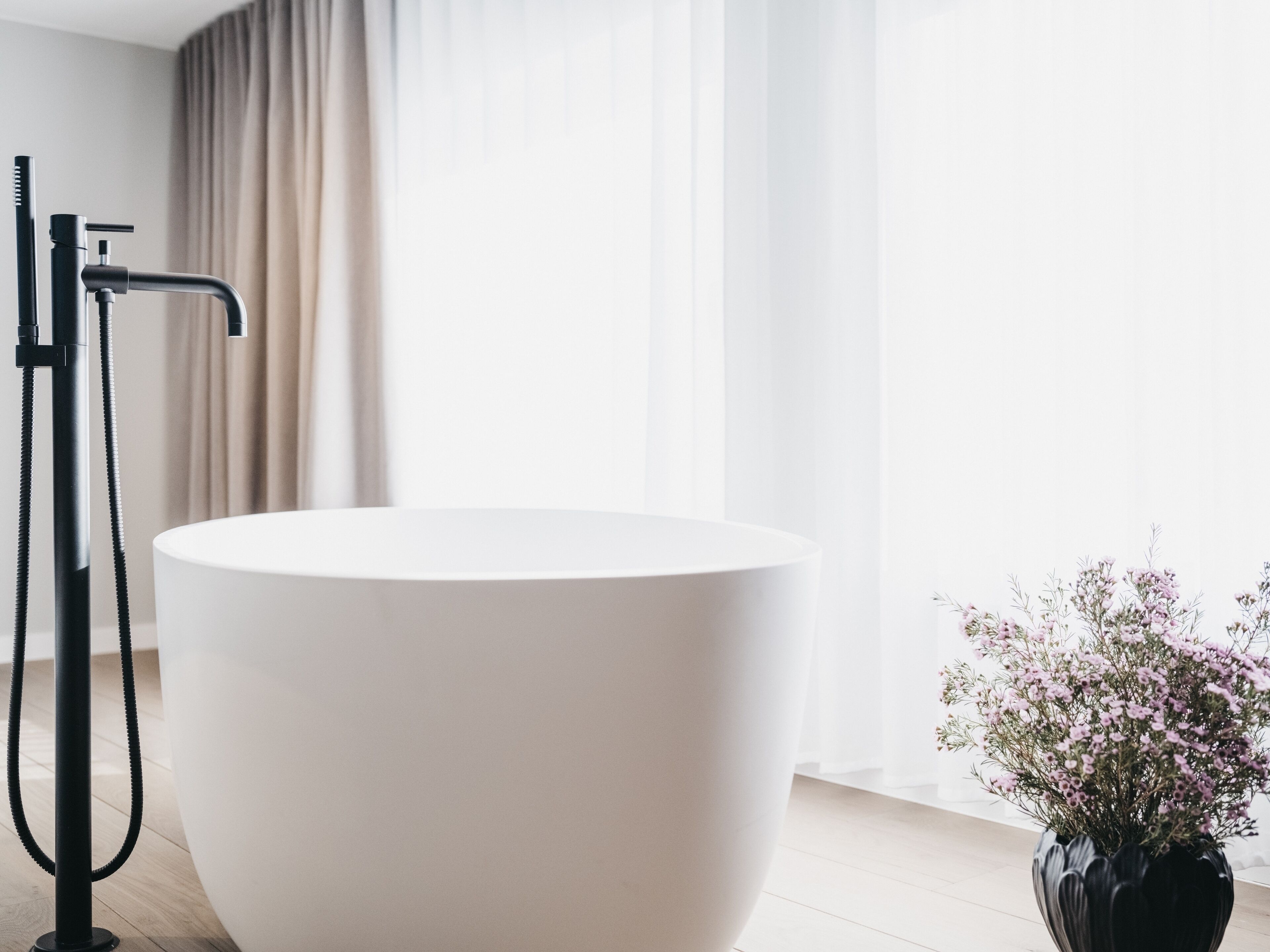 Ein modernes Badezimmer mit einer großen, weißen Badewanne, einem schwarzen Wasserhahn und einer Vase mit rosa Blumen.