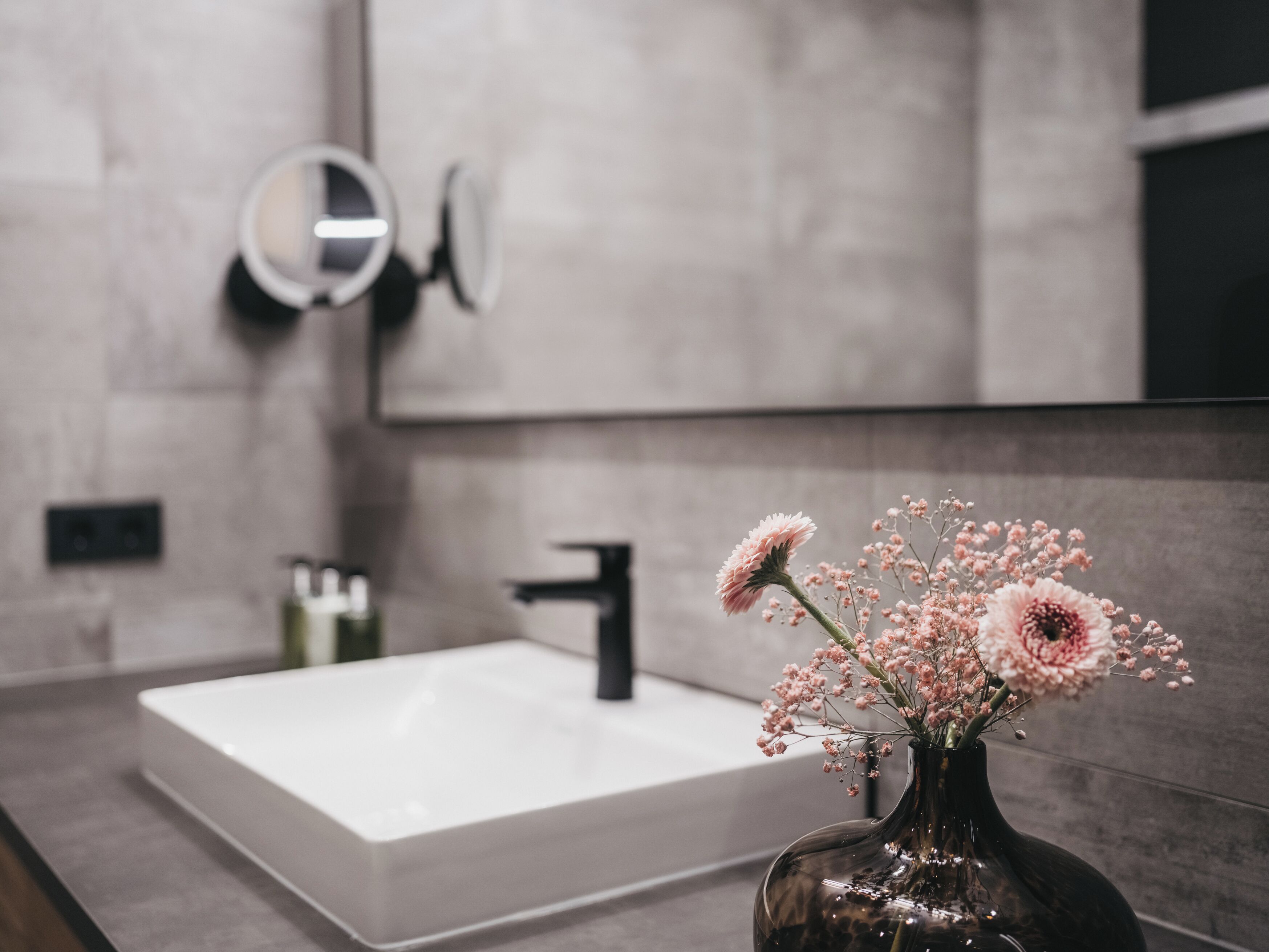 Un baño con un lavamanos blanco, un espejo redondo y un florero con flores.