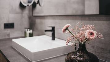 Un baño con un lavamanos blanco, un espejo redondo y un florero con flores.
