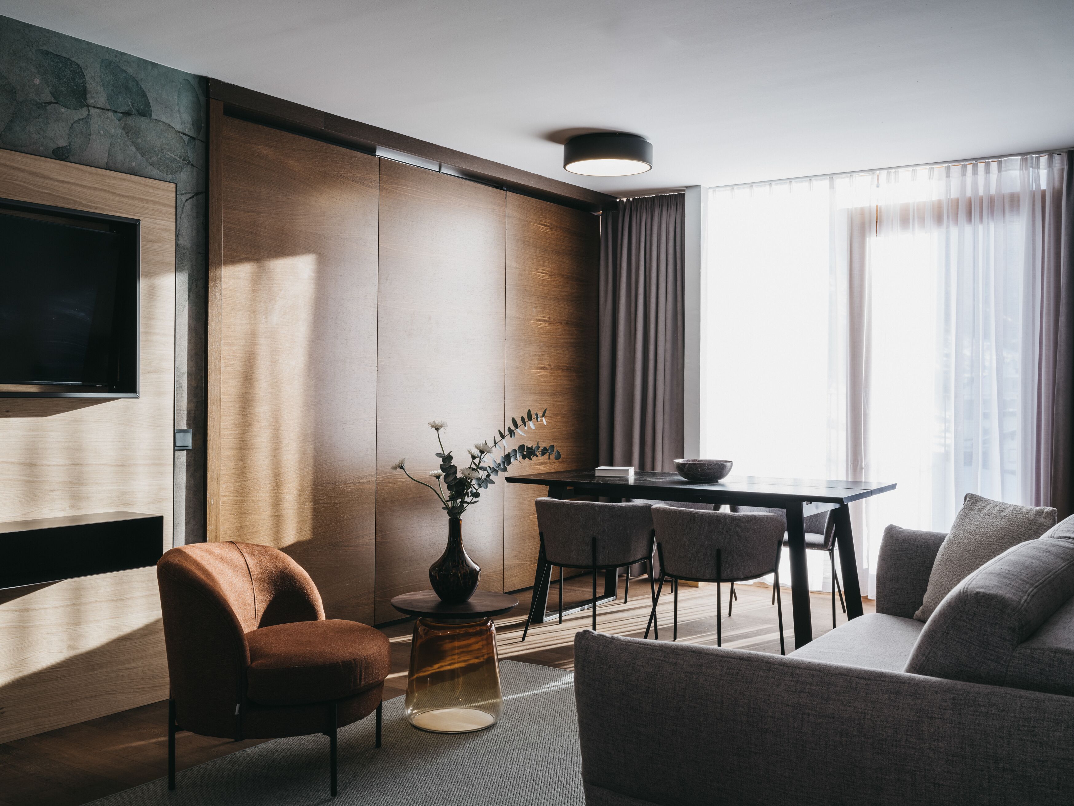 A modern living room with a flat-screen TV, a sofa, a dining table with chairs, and a vase with flowers.