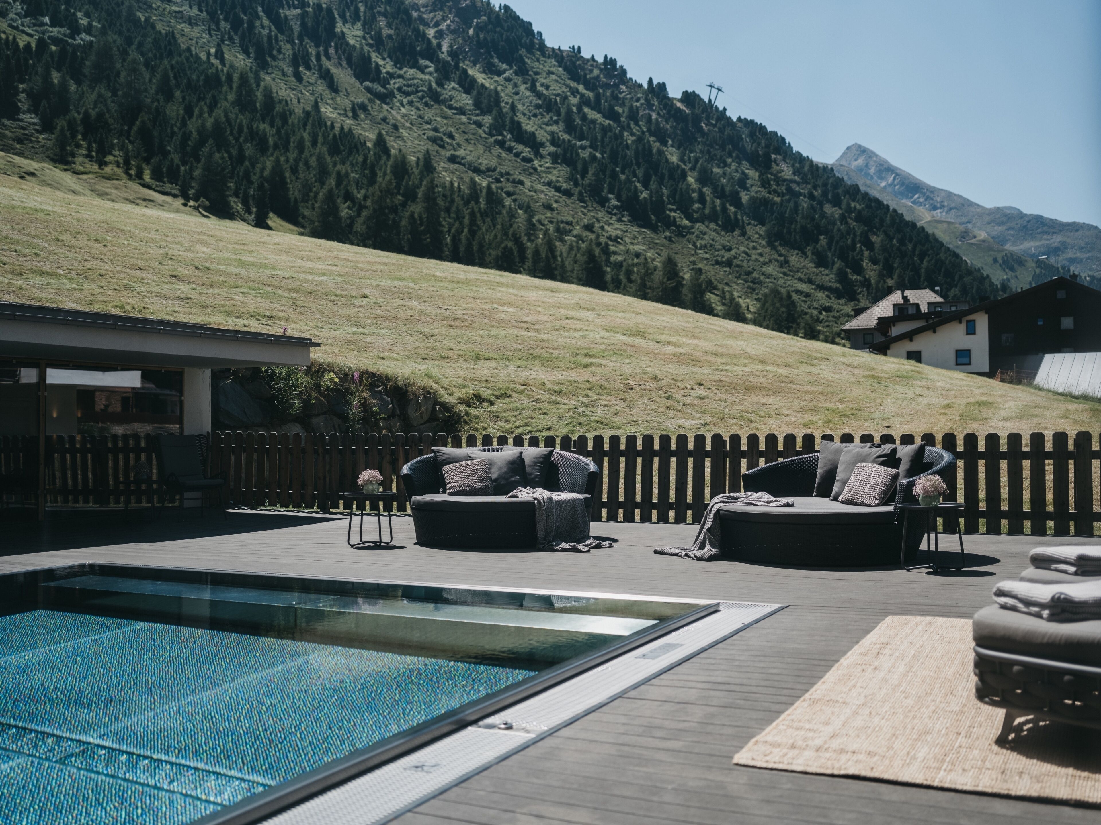 Uma área na beira da piscina com deck de madeira, dois sofás escuros com almofadas, uma mesa pequena com um vaso, e vista para montanhas e casas ao fundo.