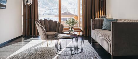 A modern hotel room with a large window offering a mountain view, a comfortable sofa, a stylish armchair, and a small round table with a vase of flowers.