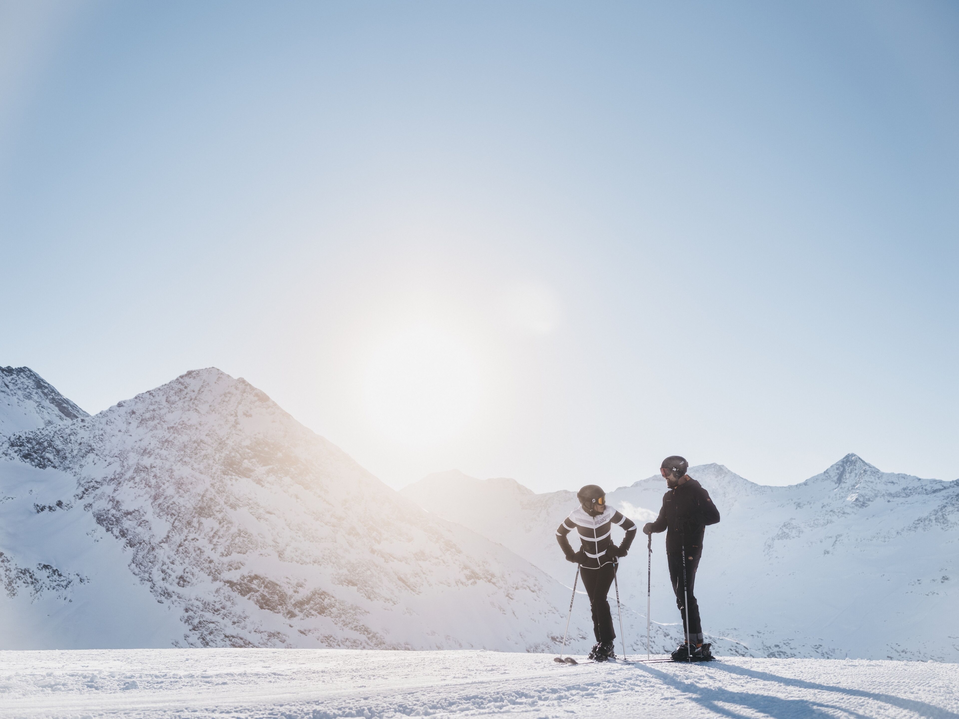 A snowy mountain landscape with two skiers standing on flat terrain.