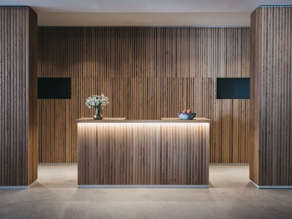 Un mostrador de recepción con un panel de madera de fondo, un jarrón con flores y un frutero.