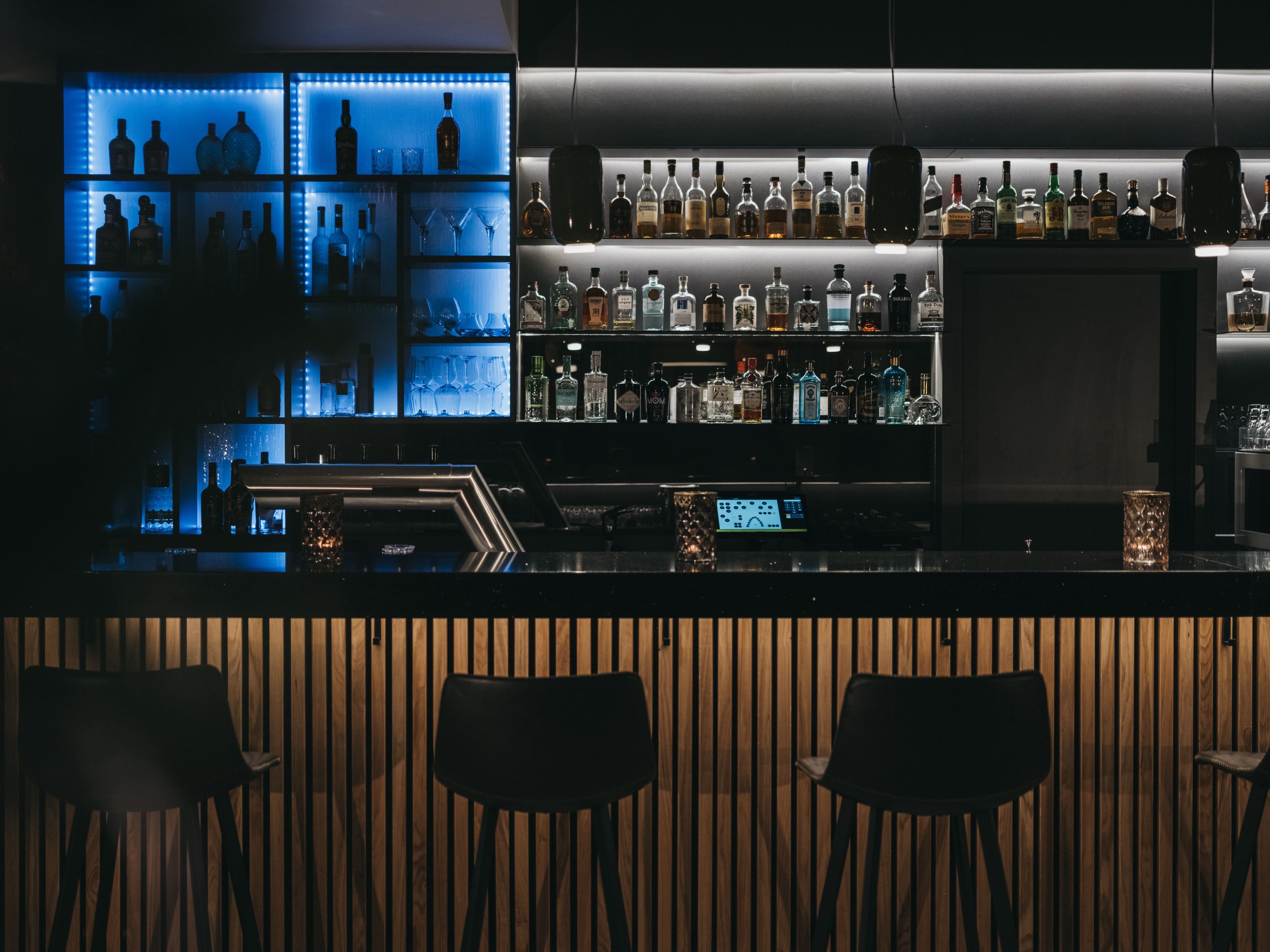 A bar area with multiple shelves of liquor bottles, a digital control panel, and three black chairs.