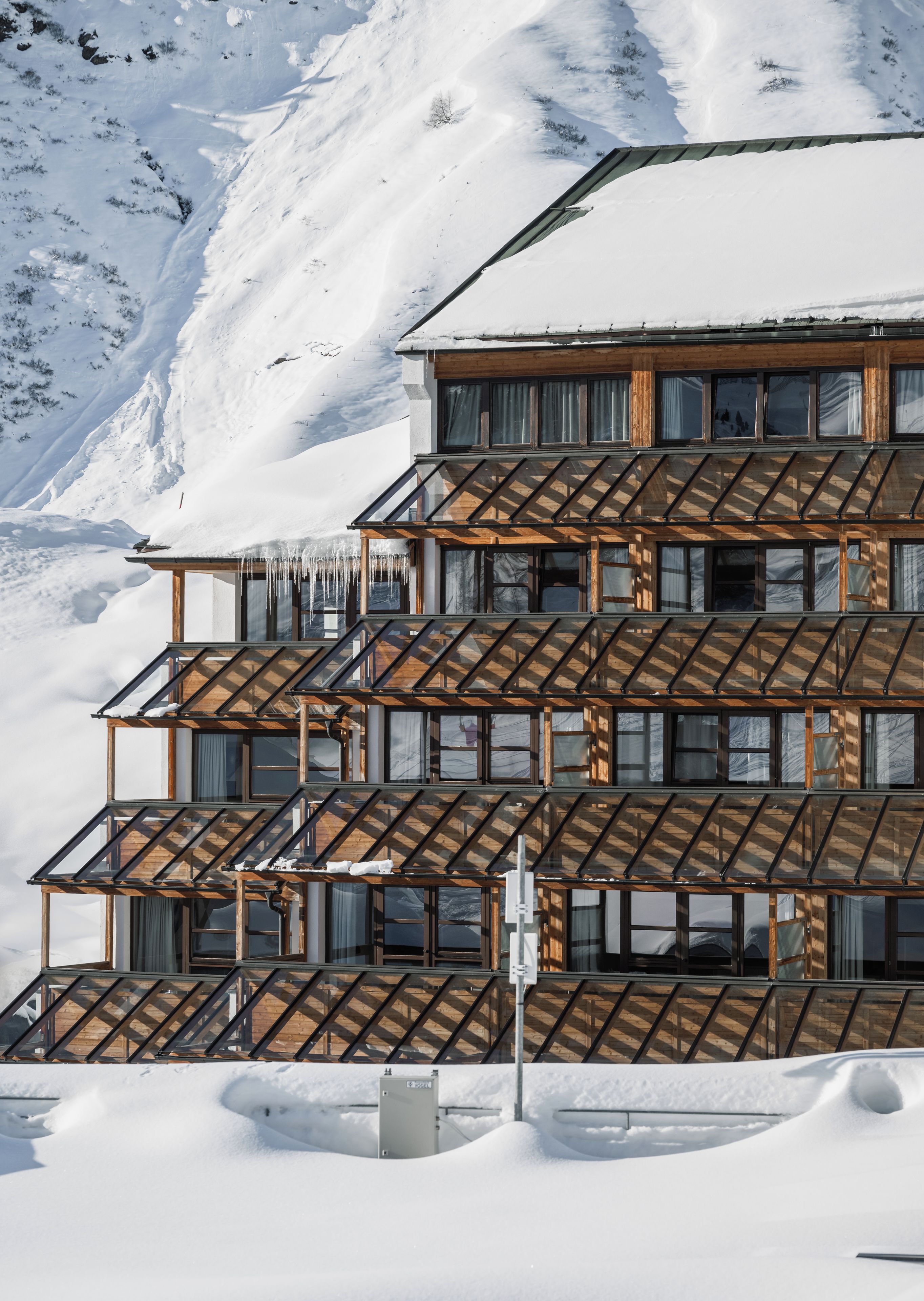 Un edificio de varios pisos con fachada de madera y grandes ventanales, rodeado de montañas nevadas.