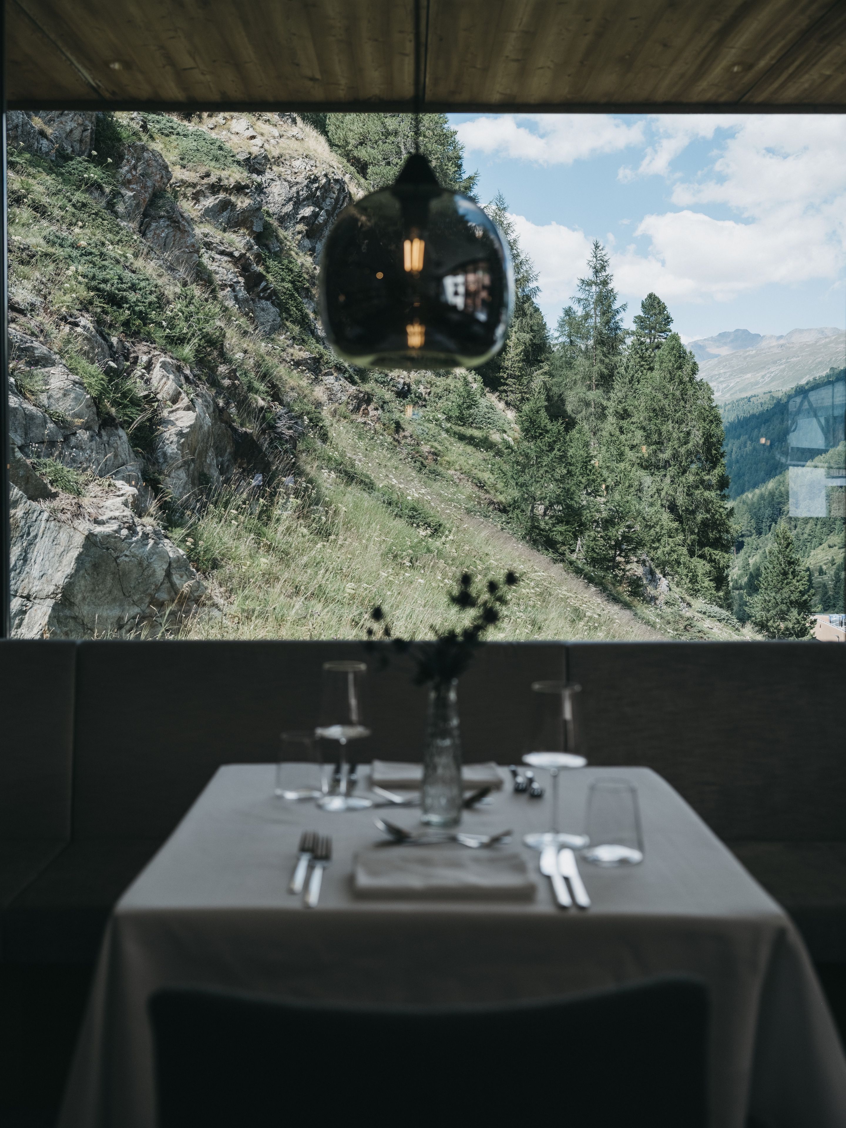 A dining table set with cutlery and a vase, with a view of a mountainous landscape through a window.