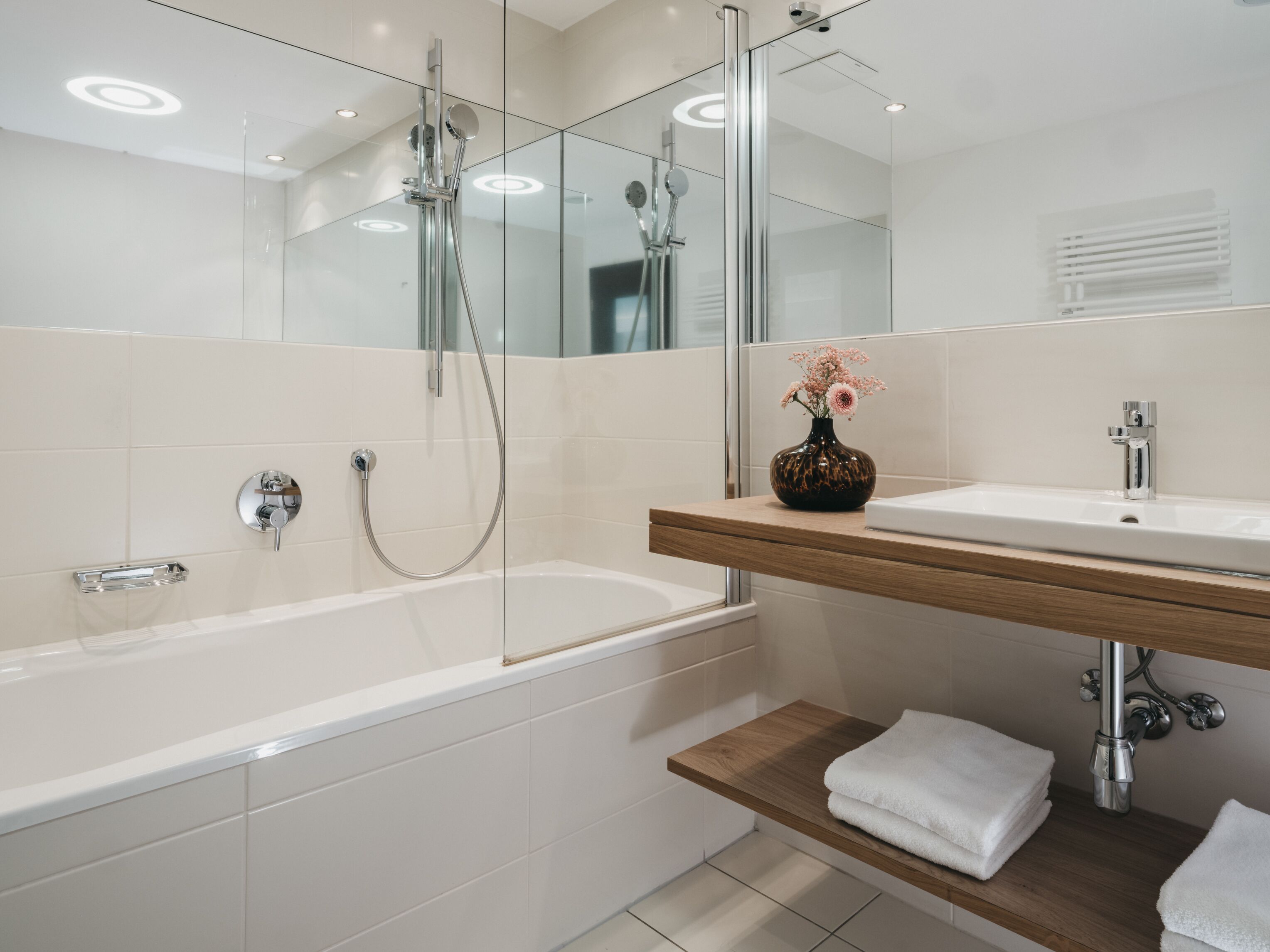 A modern bathroom with a glass-enclosed shower, a white bathtub, and a wooden vanity with a mirror.