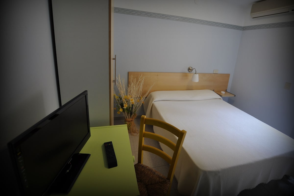 Une chambre d’hôtel comprenant un lit, un bureau avec un ordinateur portable et une télécommande, une chaise et une table de chevet avec une lampe.