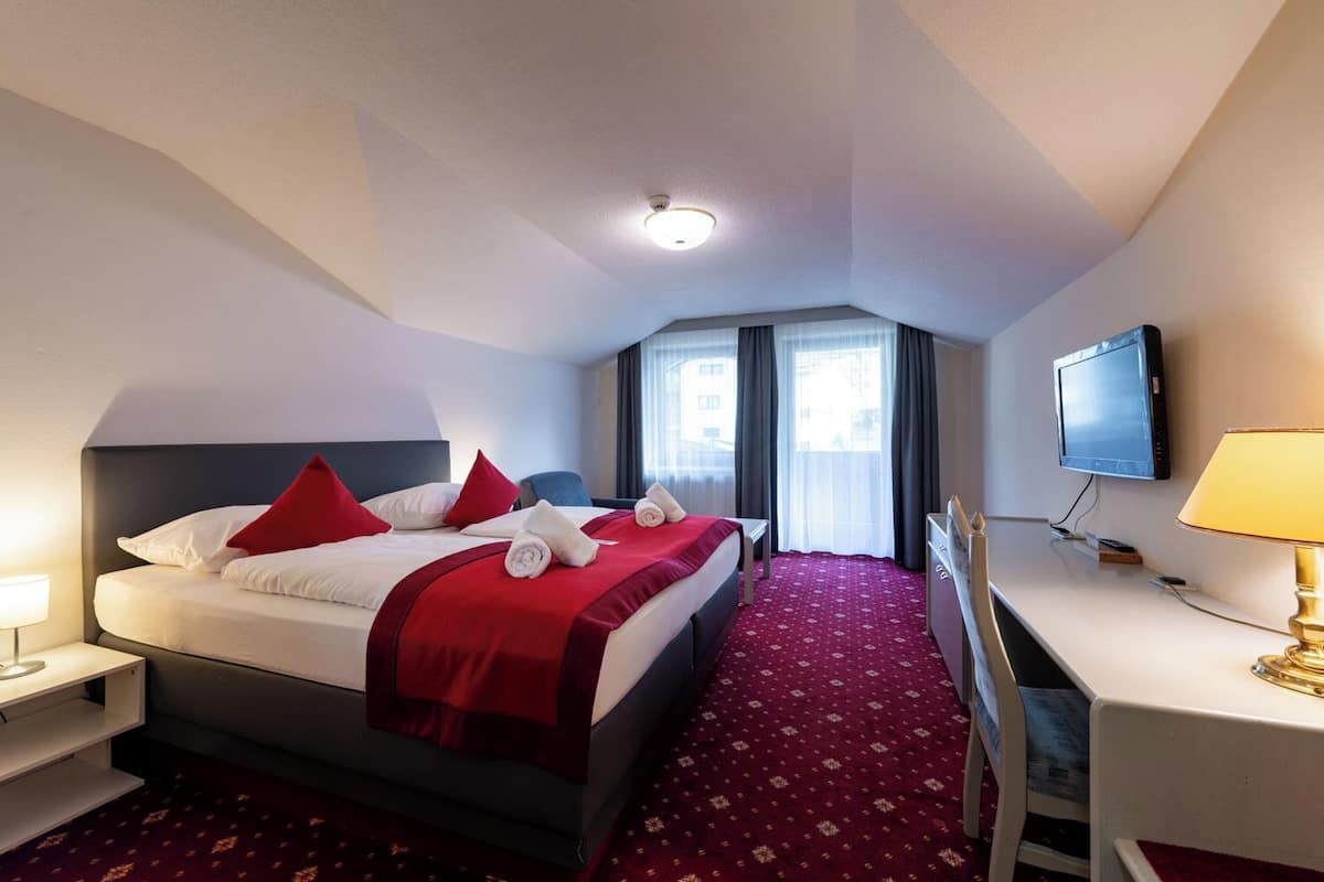 Una habitación de hotel con cama, escritorio, televisor y ventana con cortinas.