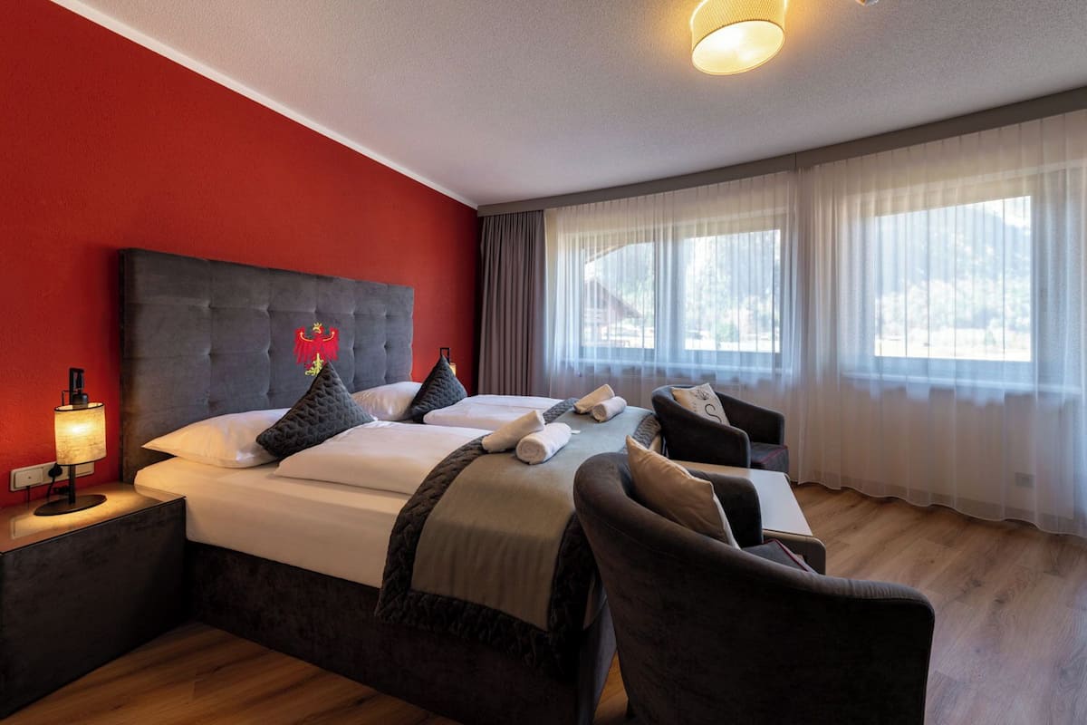 Habitación de hotel con una pared de acento roja, un cabecero capitonado, dos camas con cabeceros oscuros, una mesita de noche con lámpara, una silla y un ventanal con cortinas traslúcidas.