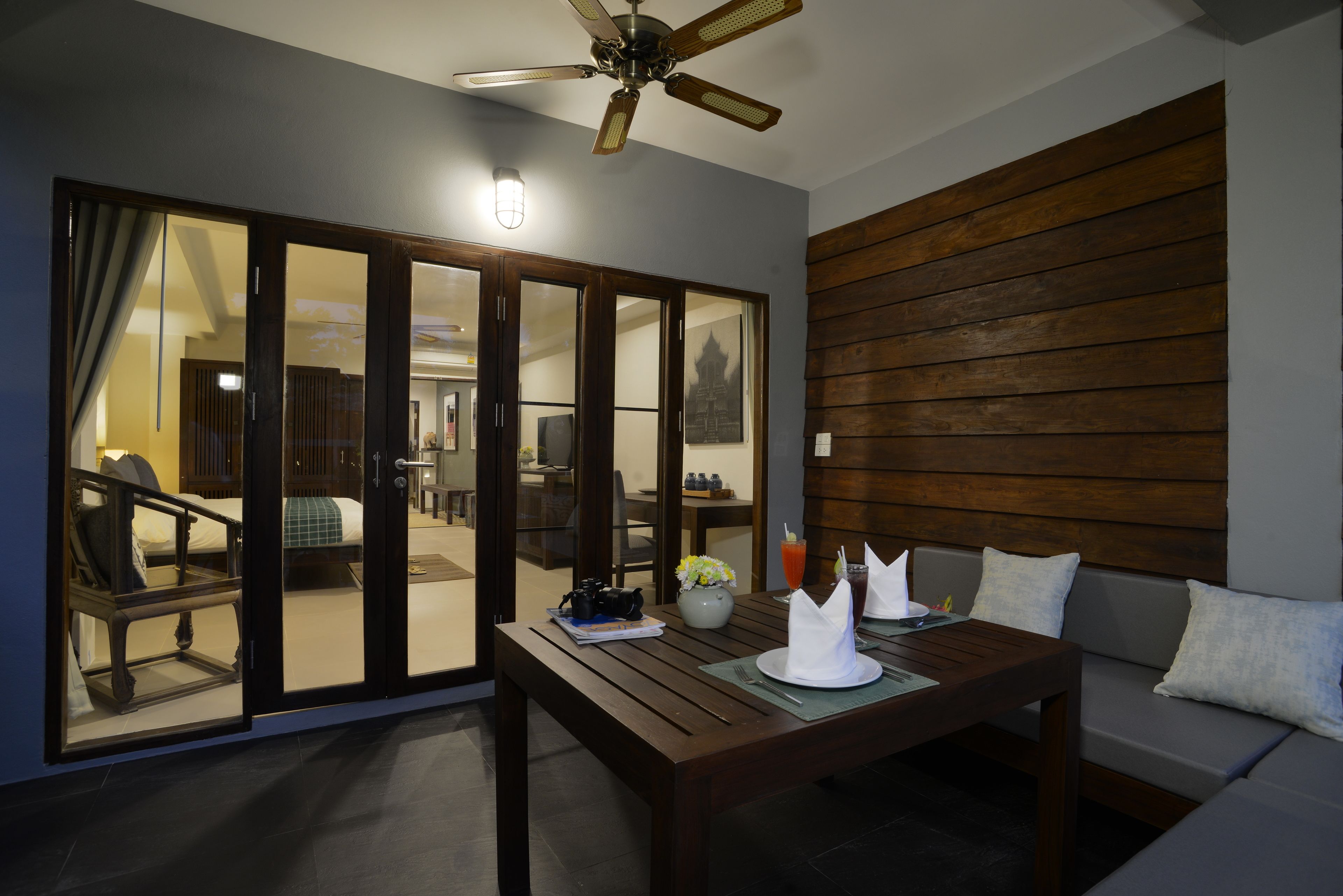 A room with a wooden ceiling fan, a dining table with a vase and napkins, a sofa with cushions, and a sliding glass door leading to another room.