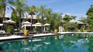 Uma área à beira da piscina com espreguiçadeiras e guarda-sóis, rodeada por palmeiras e um céu azul límpido.