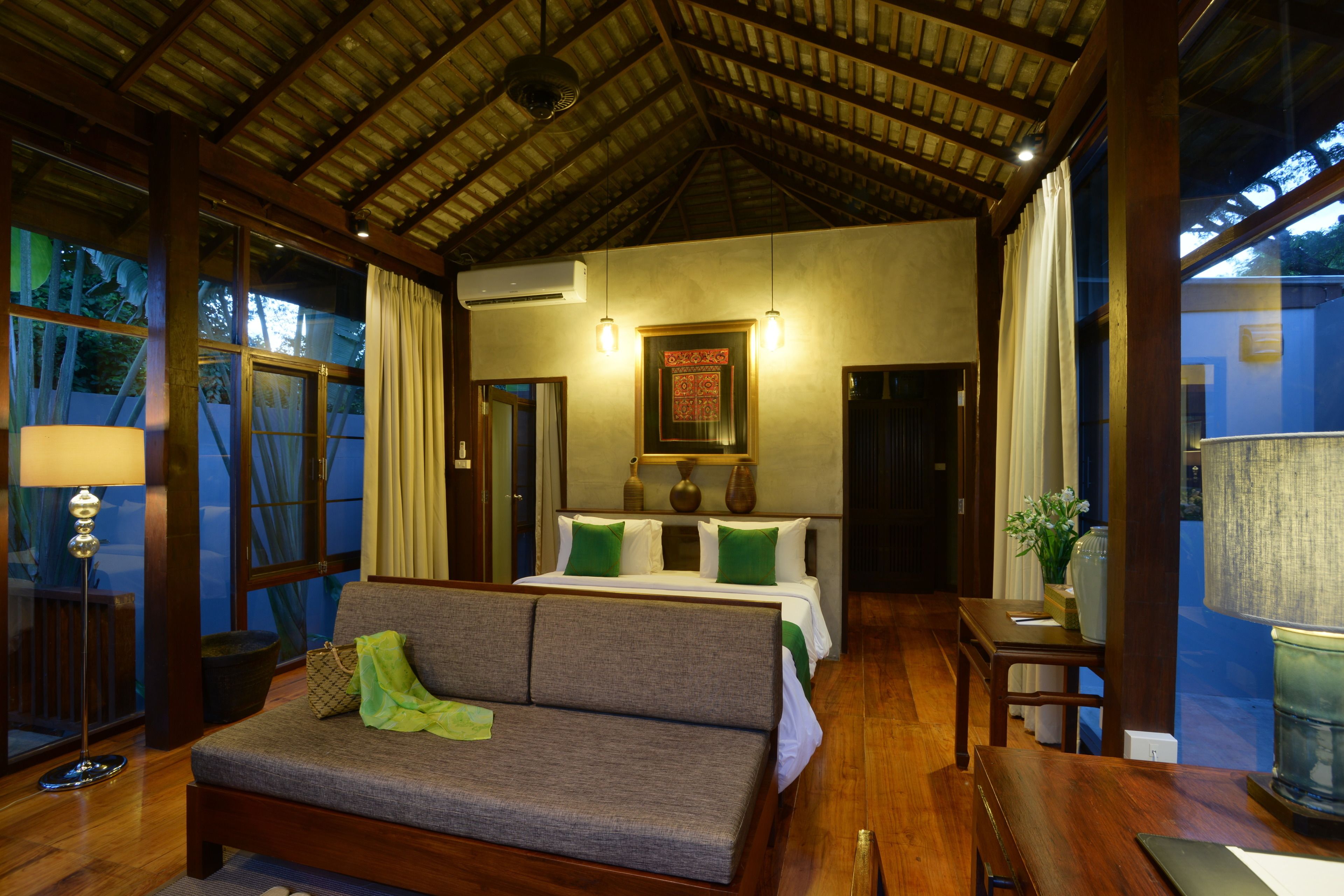 Habitación de hotel con techo de madera, sofá, cama con almohadas verdes y mesita de noche con lámpara.